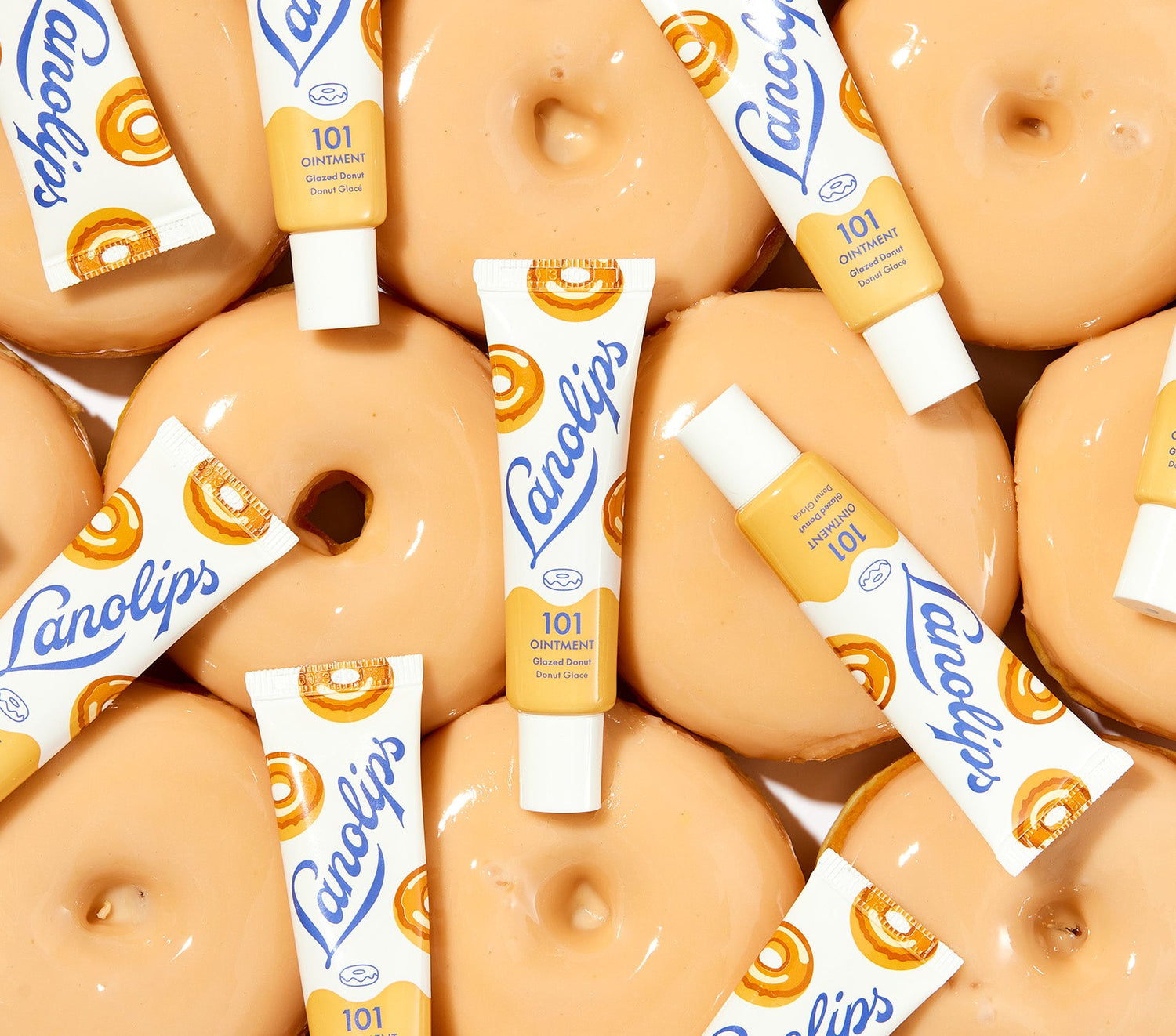 A flat lay composition featuring tubes of Lanolips 101 Ointment in the Glazed Donut flavour, placed atop freshly glazed donuts with a golden-brown hue and a glossy finish. The product contains lanolin and vitamin e, known for its deeply hydrating and nourishing properties, alongside natural oils and waxes to lock in moisture and provide long-lasting hydration.