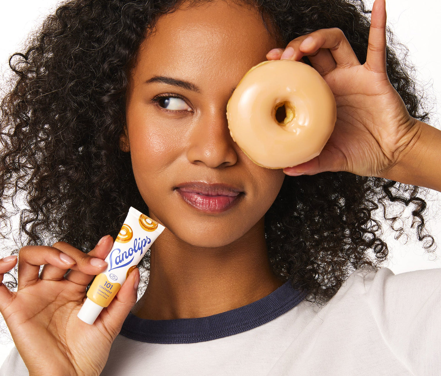Model holding a glazed donut up to her right eye while smiling. In her left hand, she holds a tube of Lanolips 101 Ointment Multi Balm in Glazed Donut. The tube features a soft yellow and white design, with blue Lanolips logo branding and illustrations of donuts. The product contains Aussie lanolin, a deeply hydrating and nourishing ingriedient that mimics the skin's natural oils. It also locks in moisture and provides a glossy, long lasting finish.