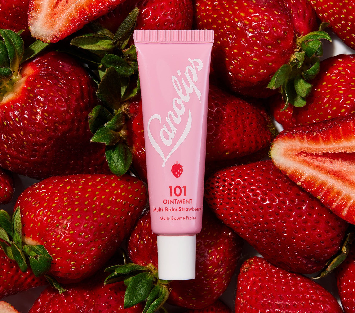 A tube of Lanolips 101 Ointment Multi-Balm Strawberry, in soft pink packaging, placed alongside fresh whole and sliced strawberries .. Made with ultra-pure lanolin, natural strawberry extract, and vitamin E, this multipurpose balm provides intense hydration and a hint of natural strawberry fruit extract, enhancing the balm's sweetness.