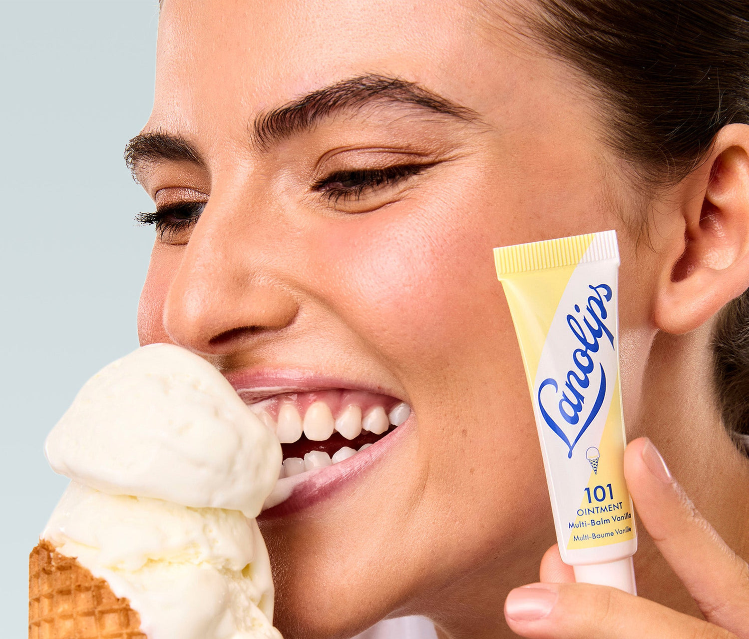 A close up shot of a model holding a tube of Lanolips 101 Ointment Multi Balm in Vanilla close to her face, while eating a vanilla ice cream cone. The balm's soft yellow packaging with the Lanolips blue lettering and an ice cream illustration. Made with lanolin, vitamin e and natural vanilla extract, this super rich balm provides intense hydration served in a sweet, dessert-like scent.