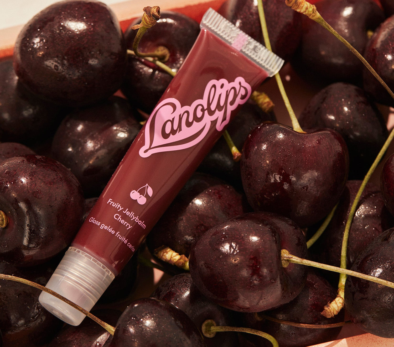 A tube of Lanolips Fruity Jellybalm in Cherry, nestled among fresh, glossy cherries. The rich red packaging features a pink Lanolips logo and a cherry icon, reflecting its juicy, fruit-inspired formula. Made with ultra rich lanolin and all natural cherry extract, this balm provides deep moisture and a sheer, glossy tint, inspired by the sweetness of ripe cherries.