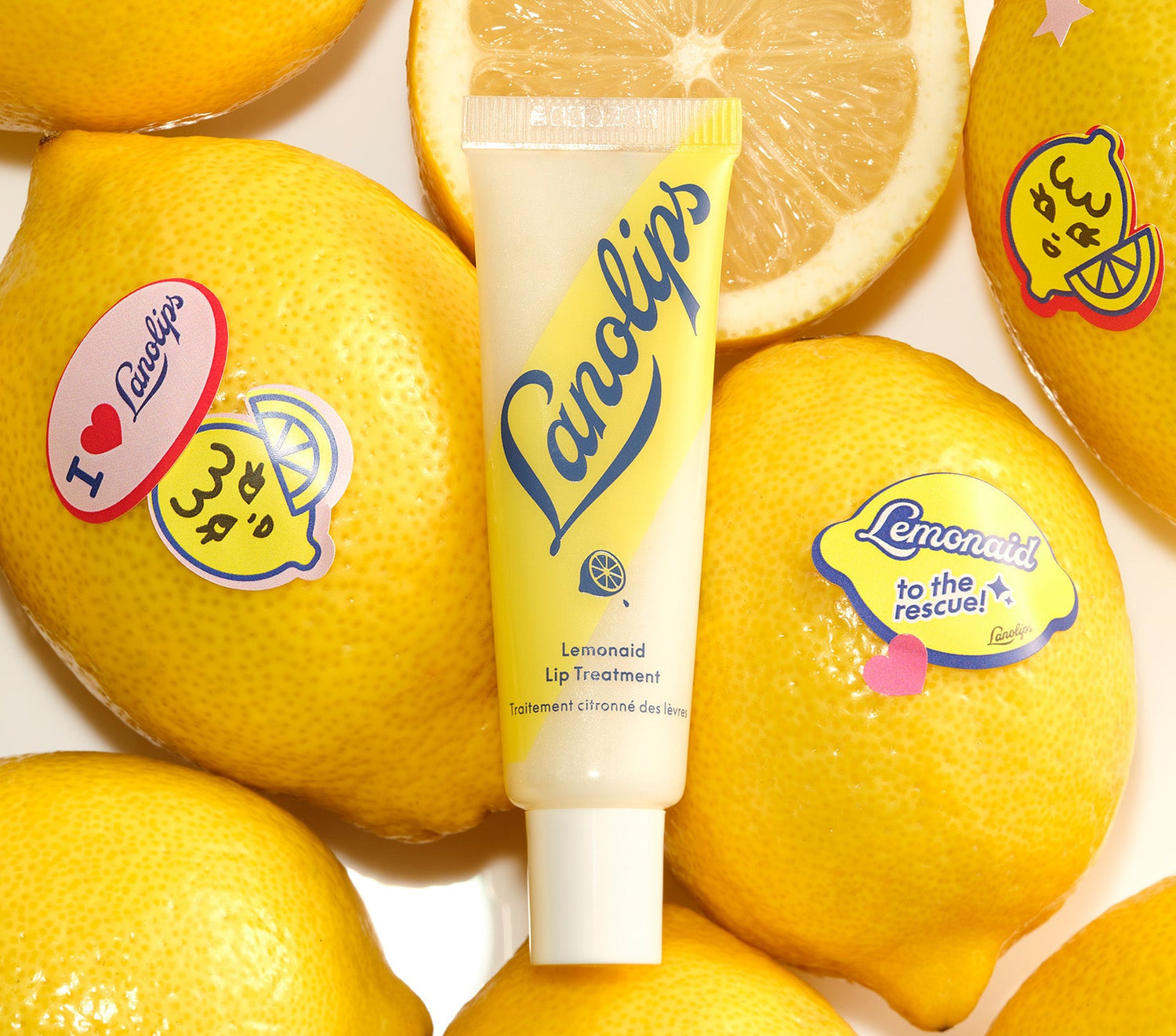 A tube of Lanolips Lemonaid Lip Treatment sits among fresh, bright yellow lemons, some decorated with playful stickers. The packaging features a yellow and white gradient with bold blue Lanolips lettering and a small lemon icon. The lip treatment, infused with rich lanolin, all-natural lemon oil, vitamin e and a hint of shimmer, deeply nourishes, gently exfoliates, and leaves lips feeling soft, smooth, and hydrated with a glossy finish.