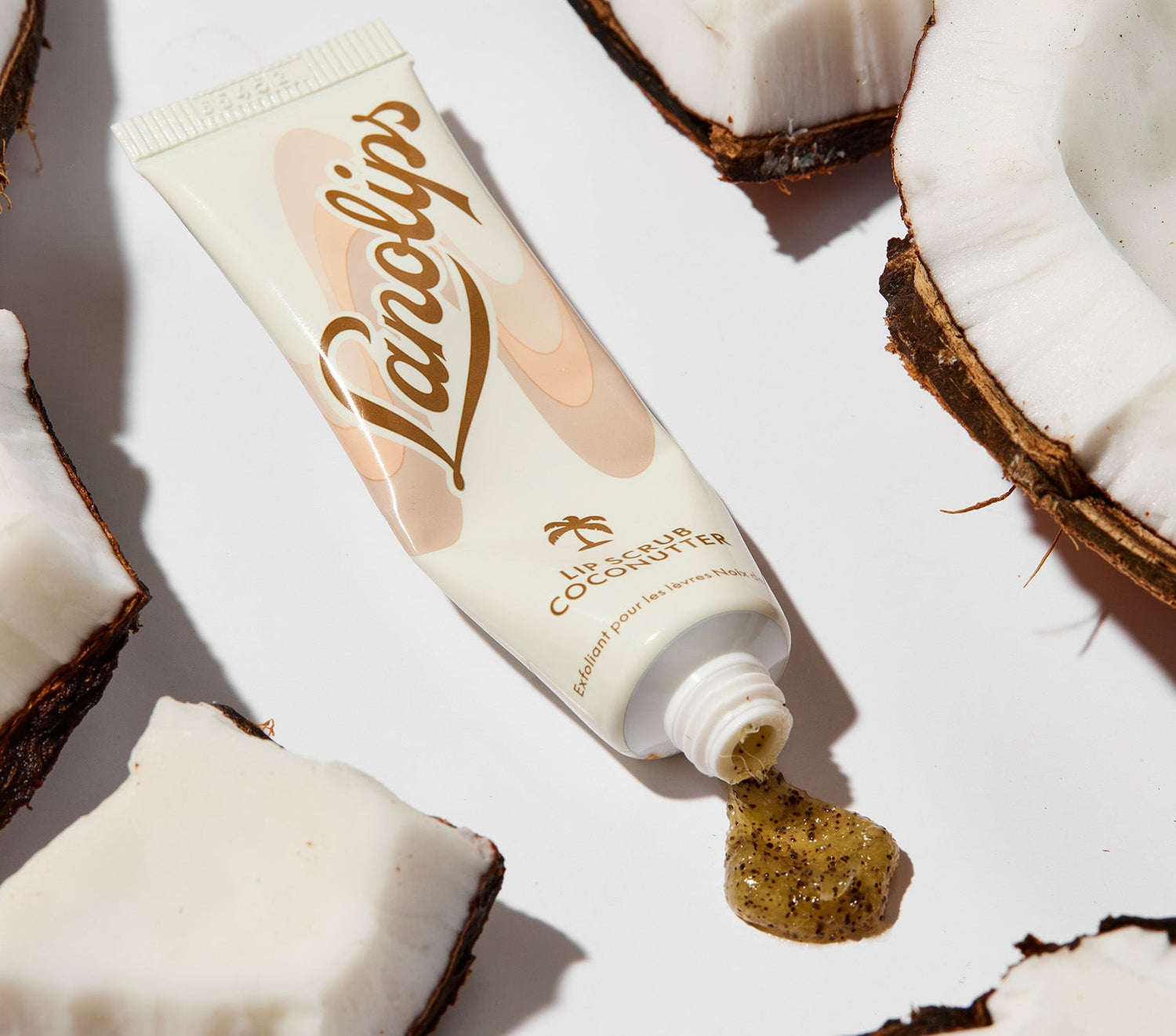 A tube of Lanolips Lip Scrub in Coconutter rests among cracked coconut pieces, with its exfoliating formula spilling out. The packaging features a sleek beige and brown design with the Lanolips logo and a small palm tree icon. This 100% natural balm-based scrub is made with gentle sugar and real, finely ground coconut shell pieces to smooth away dry, flaky skin. Infused with ultra-pure lanolin and coconut oil, it deeply nourishes while gently buffing away dead skin, leaving lips soft, smooth, and hydrated.