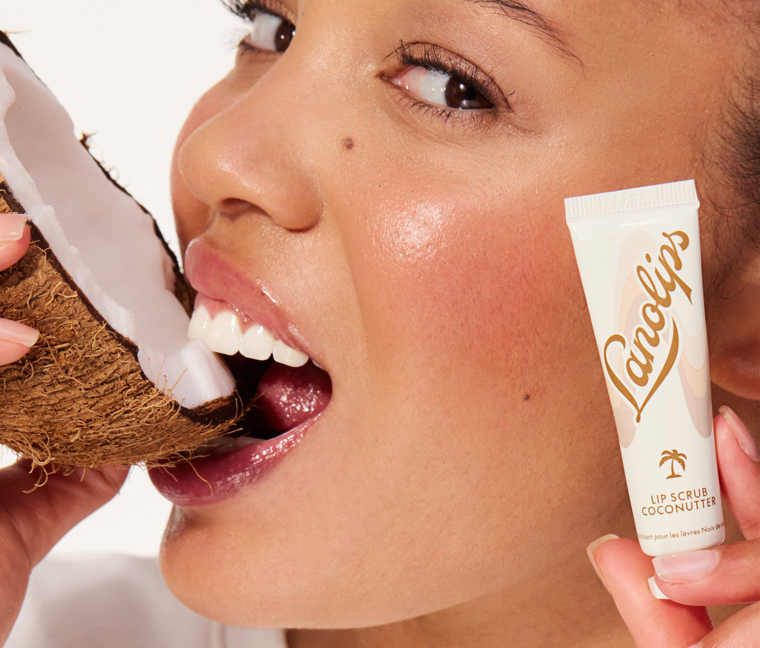 A model holds a cracked coconut close to her lips while showcasing a tube of Lanolips Lip Scrub in Coconutter. The packaging features a sleek beige and brown design with the Lanolips logo and a small palm tree icon. This 100% natural balm-based scrub is made with gentle sugar and real, finely ground coconut shell pieces to smooth away dry, flaky skin. Infused with ultra-pure lanolin and coconut oil, it deeply nourishes while gently buffing away dead skin, leaving lips soft, smooth and hydrated.