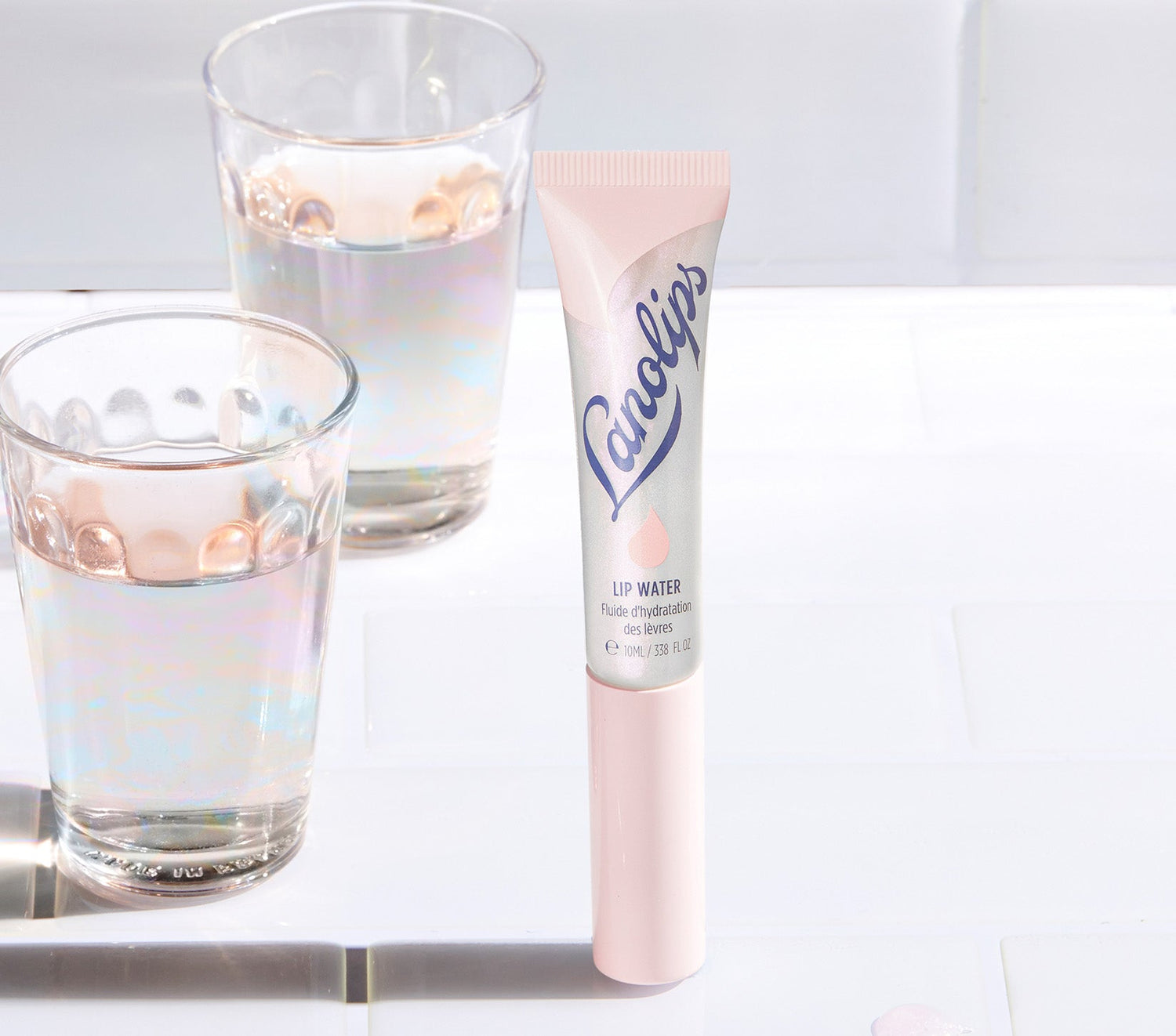 A tube of Lanolips Liquid Lip Water stands on a white surface, with shimmering water glasses in the background. The silver and pink packaging features the blue Lanolips branding and a droplet icon. Made with liquid lanolin, hyaluronic acid, glycerin and peppermint, this non-sticky, hydrating lip serum delivers deep moisture with a underwater holographic glow. Like a drink of water for your lips, it nourishes, softens and adds a glass like sheen - perfect for dry lips and lip lickers.