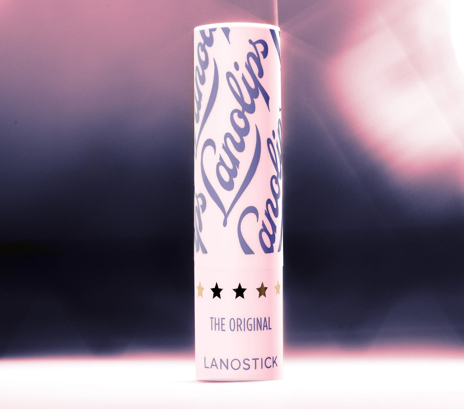 A sleek tube of Lanolips Lanostick stands against a soft pink and black gradient background. This 90s-inspired wind-up lip balm is perfect for pockets, bags, and on-the-go hydration. From the makers of the 'World’s Best Lip Balm,' Lanostick glides on smoothly, delivering a rich layer of moisture that deeply conditions lips. Infused with lanolin, natural waxes, butters, and vitamin E, it nourishes and locks in hydration for hours. The ultimate day-balm for soft, healthy lips.