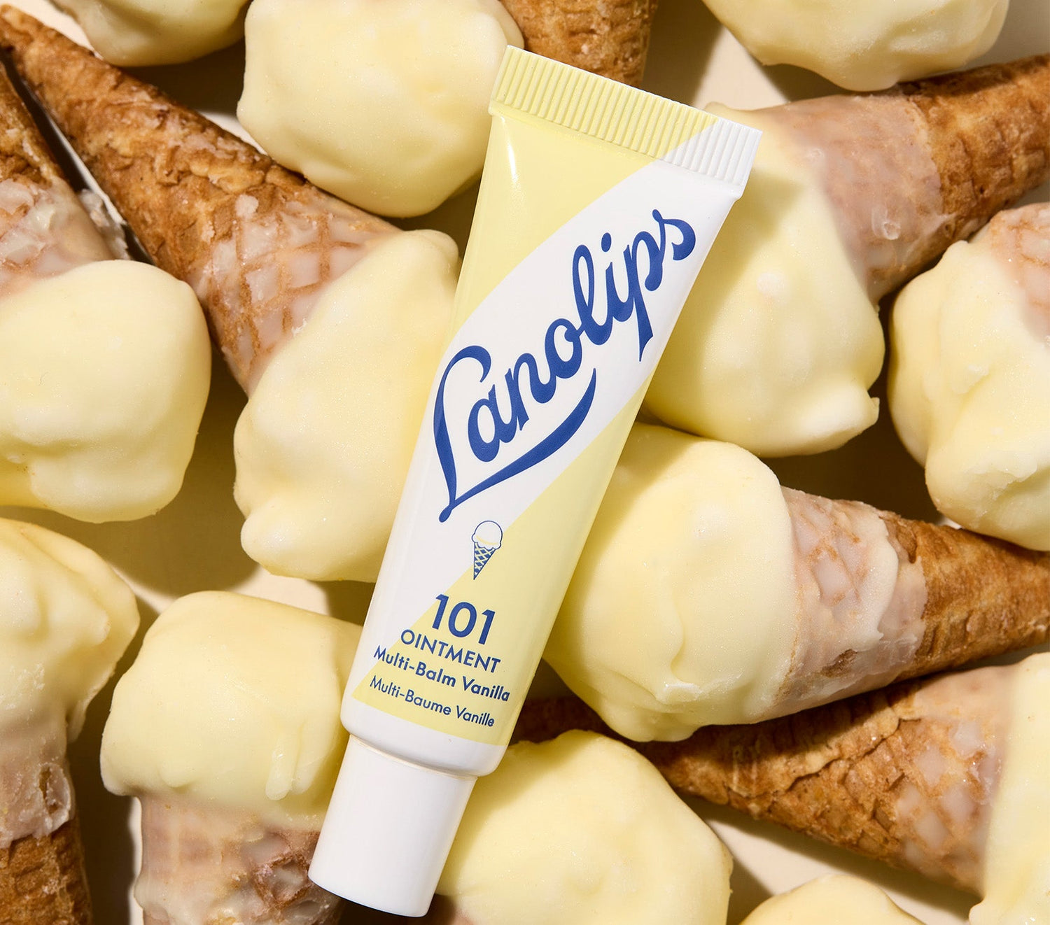 A tube of Lanolips 101 Ointment Multi-Balm in Vanilla, placed on top of small waffle cones dipped in creamy vanilla. The balm's packaging is soft yellow with blue lettering, featuring an ice cream cone illustration. Made with lanolin, vitamin e, and vanilla extract, this super rich balm penetrates & seals in moisture to give long-lasting hydration for dry & chapped lips, skin patches, cuticles, elbows & more.