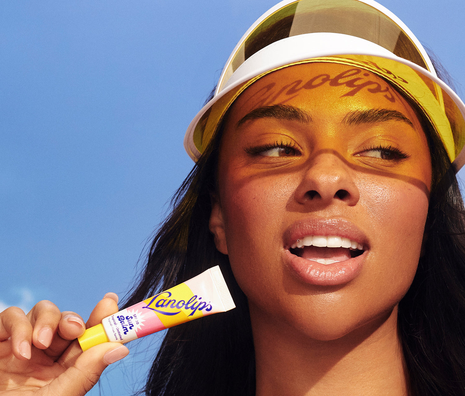 A model wears a yellow-tinted sun visor that casts the word “Lanolips” across her glowing face. The model holds a tube of Lanolips Sun Balm SPF 30 Tropical lip balm and smiles against a clear blue sky. Her glossy lips and golden makeup reflect the beachy, sun-safe, and hydrating vibe of the product - It's a sun visor for your lips.