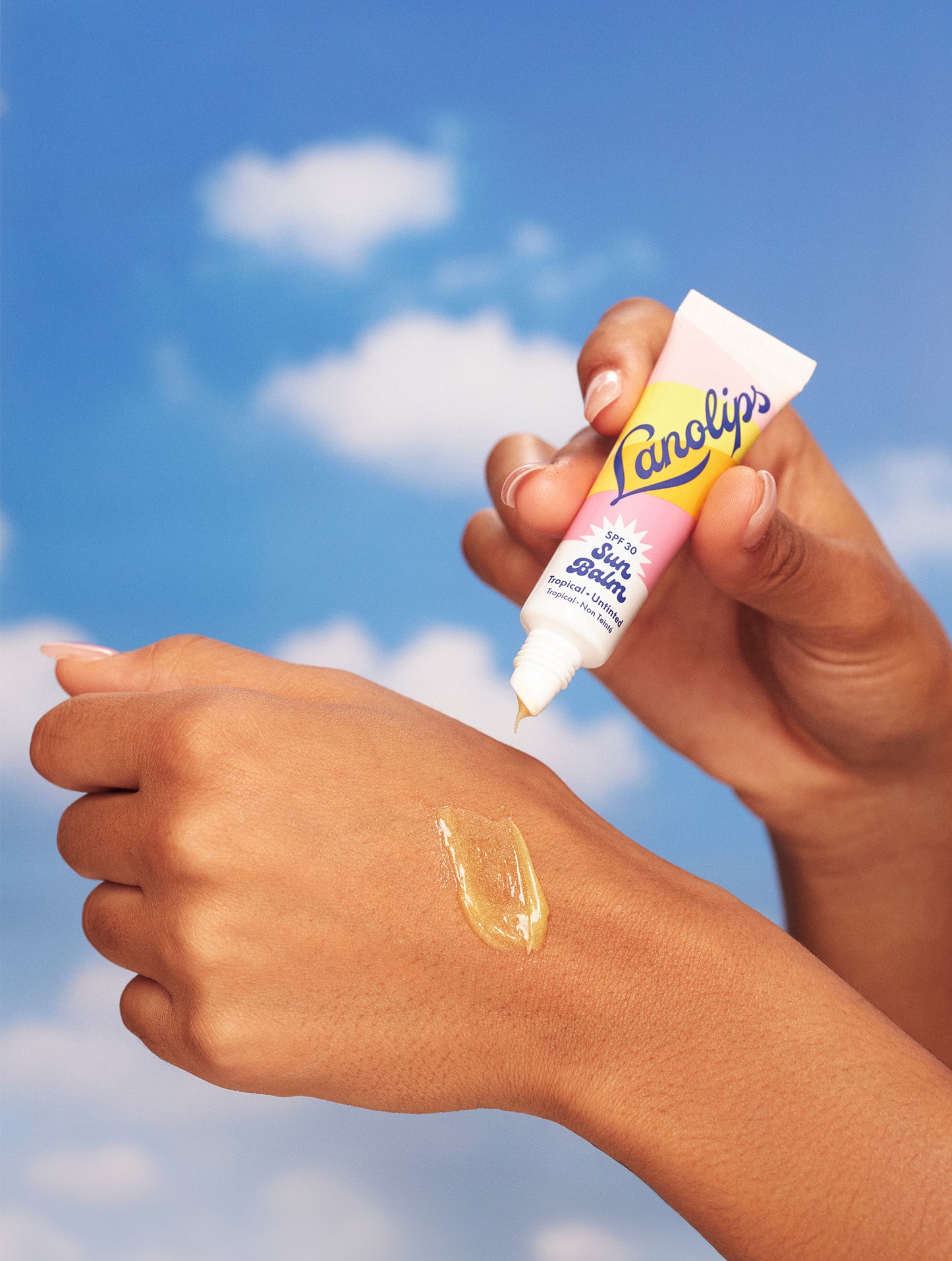 Hands apply Lanolips Sun Balm SPF 30 Tropical on skin, showing its glossy, golden texture - hydrating, protective and tropically nourishing.