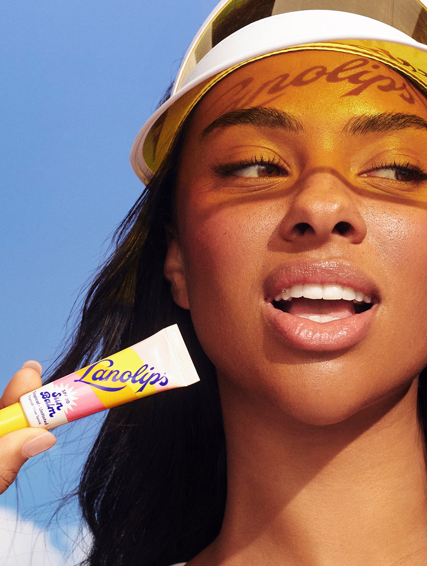 A model holds a tube of Lanolips Sun Balm SPF 30 Tropical lip balm against a blue sky backdrop. Wearing a white-and-yellow sun visor, the word “Lanolips” is cast as a warm shadow across her forehead. Her glossy lips and radiant skin reflect the balm’s protective, hydrating, and summery feel - perfectly capturing SPF-meets-skincare for everyday glow.