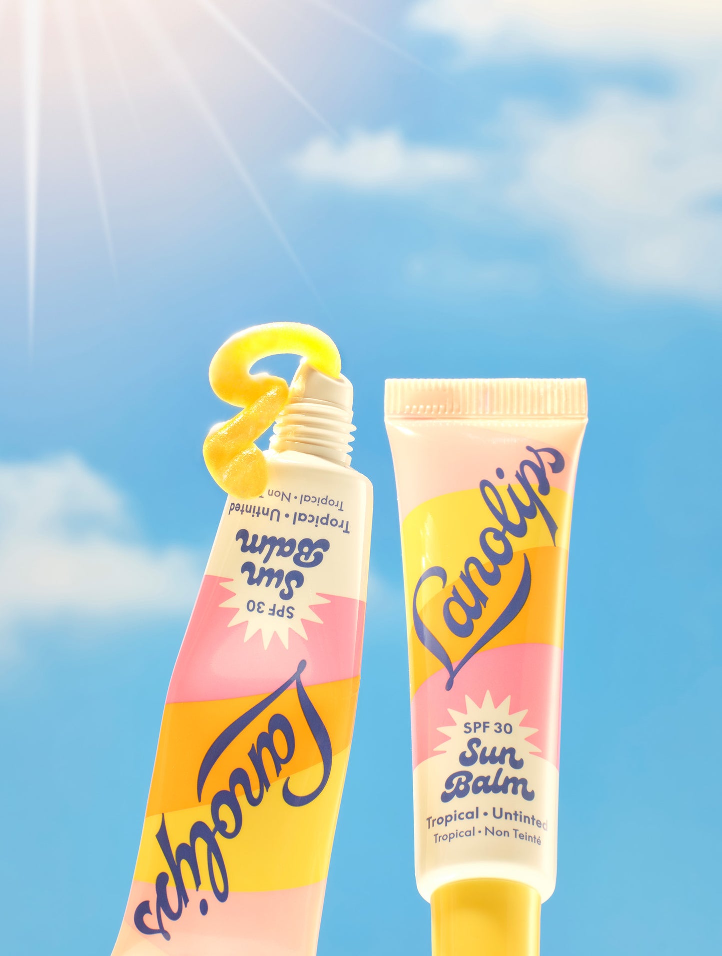 Set against a radiant blue sky with a shining sun, two tubes of Lanolips Sun Balm SPF 30 Tropical lip balm are featured - one standing upright and the other squeezed to reveal a vibrant, glossy balm shaped like a “3” to highlight its SPF 30 protection. This creamy, radiant formula is packed with hydrating lanolin, SPF protection and a luminous sheen.