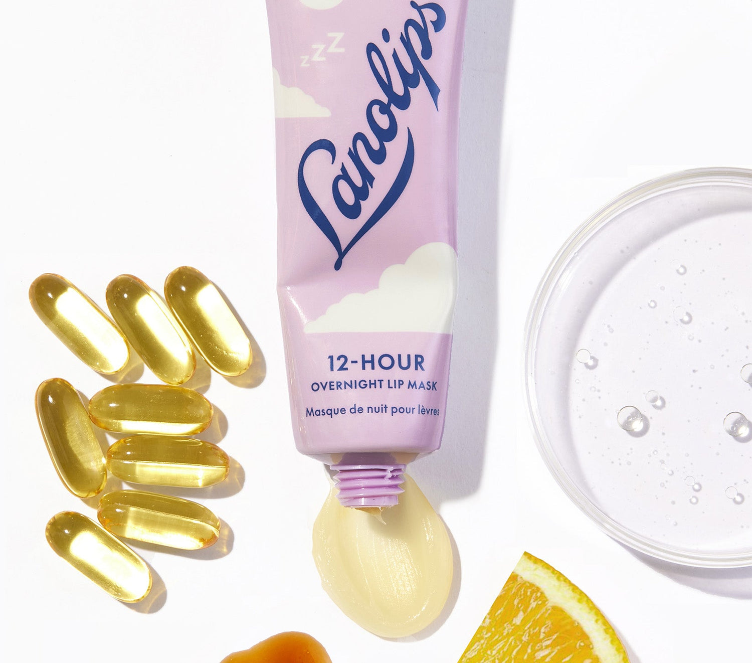 A tube of Lanolips 12 Hour Overnight Lip Mask. The tube is slightly squeezed, with a glossy creamy swipe of the lip mask visible at the opening. Surrounding the product are the key ingredients including vitamin e capsules, a glass dish containing hyaluronic acid, a drizzle of manuka honey and a fresh slice of orange, representing vitamin c. The lip mask contains ultra-pure grade lanolin, hyaluronic acid, ceramides, vitamin c, ceramides, vitamin e - to help repair and nourish dry lips by morning.