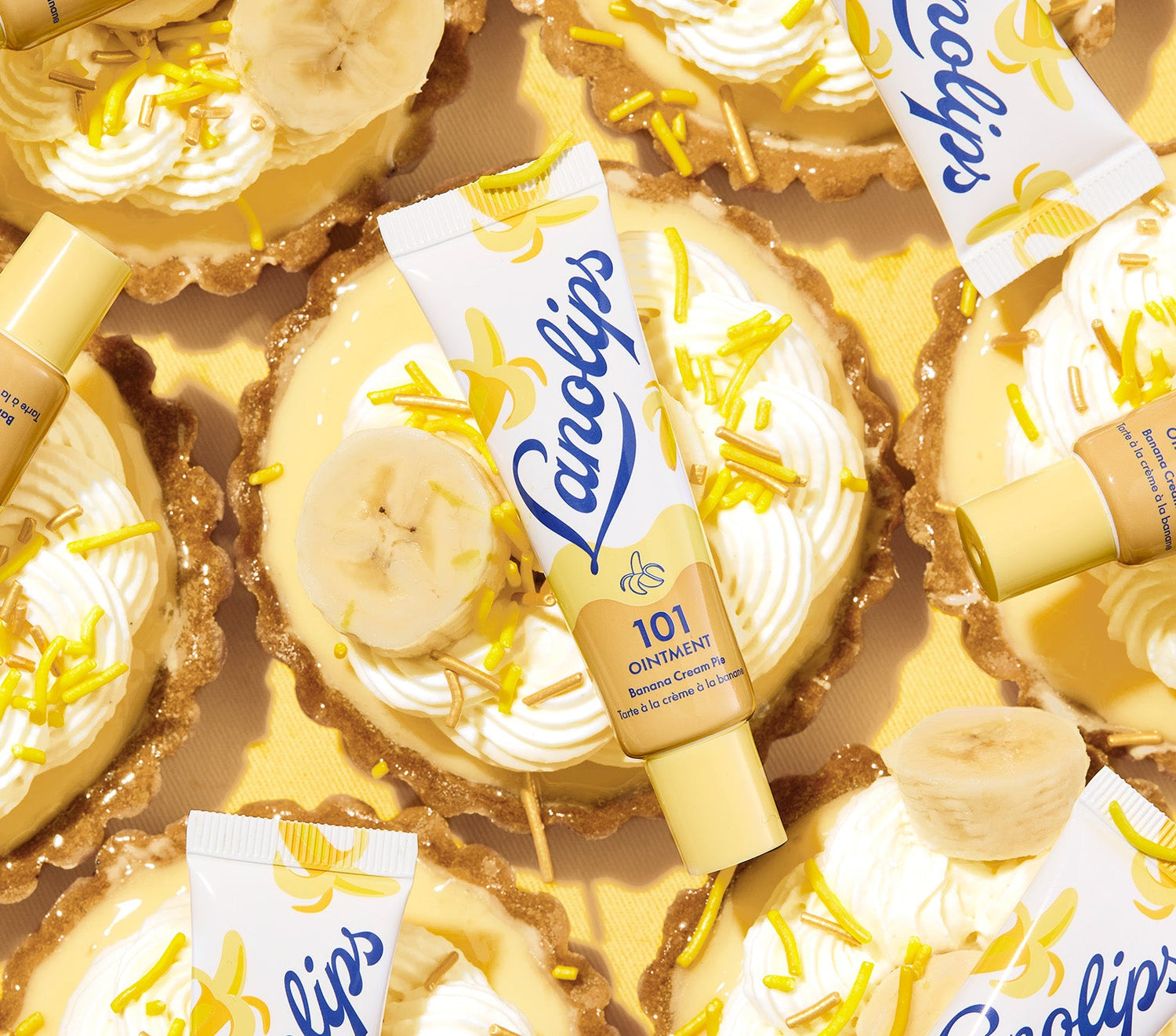 A vibrant flatlay of Lanolips 101 Ointment Multi Balm Banana Cream pie flavour, surrounded by banana cream tarts. The tarts are topped with whipped cream, banana slices, and yellow sprinkles. The packaging of the ointment features a creamy yellow design with a banana illustration, complementing the dessert aesthetic. The composition is bright and warm, with golden tones enhancing the banana theme. Made with lanolin, vitamin e and natural flavours to hydrate dry & chapped lips.