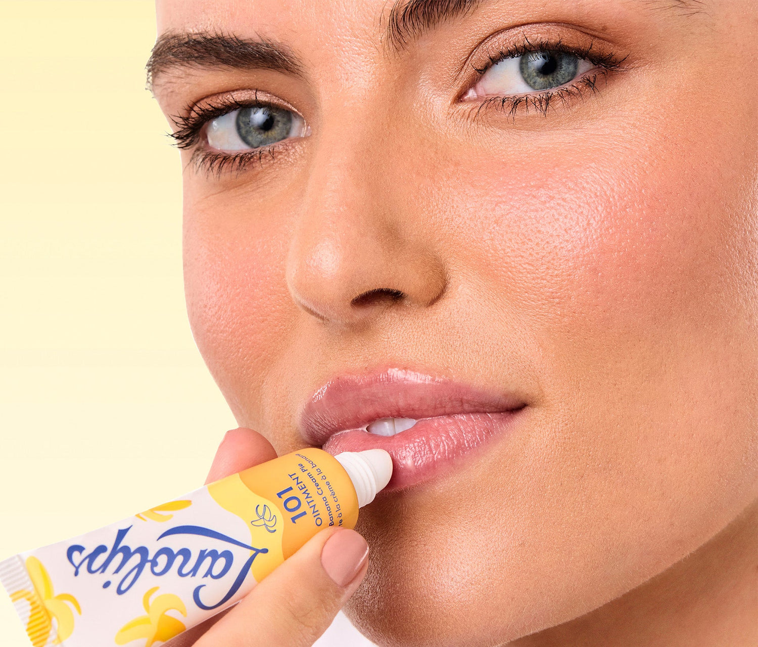 A close up shot of a model applying Lanolips 101 Ointment Multi Balm in Banana Cream Pie flavour to her lips. She holds the yellow and white tube her mouth, with applicator gently touching her lips. The background is pastel yellow. Made with lanolin, vitamin e and infused with a hint of biscuit, caramel & sweet pastry to hydrate dry & chapped lips.