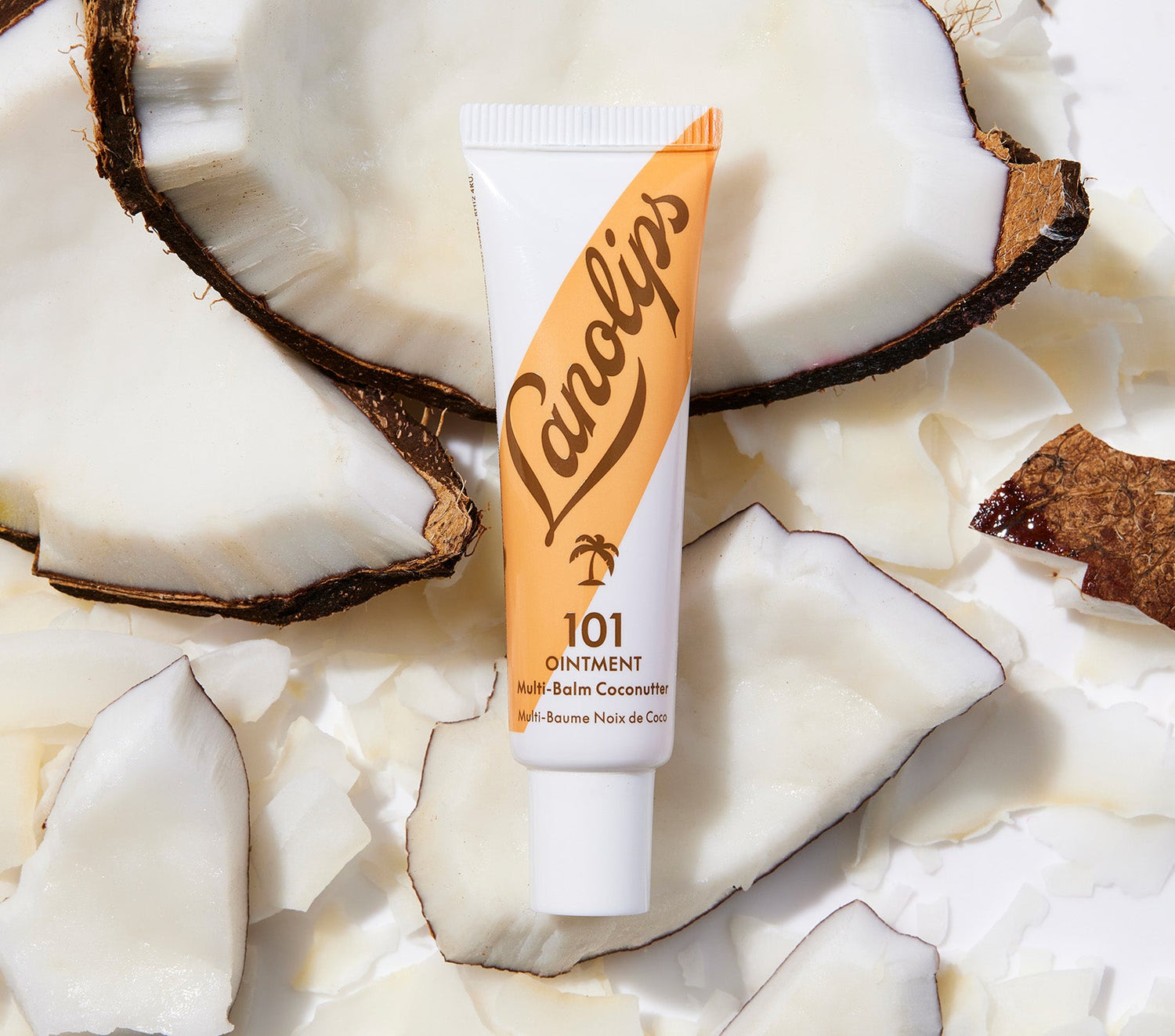 A tube of Lanolips 101 Ointment Multi Balm Coconutter laying against a background of fresh coconut pieces. The packaging features a warm caramel and white gradient design with a palm tree illustrtion, complementing the tropical coconut theme. Using ultra-pure grade Aussie lanolin, vitamin e and coconut, his super-rich balm penetrates & seals in moisture to give long-lasting hydration for dry & chapped lips, skin patches, cuticles, elbows & more.