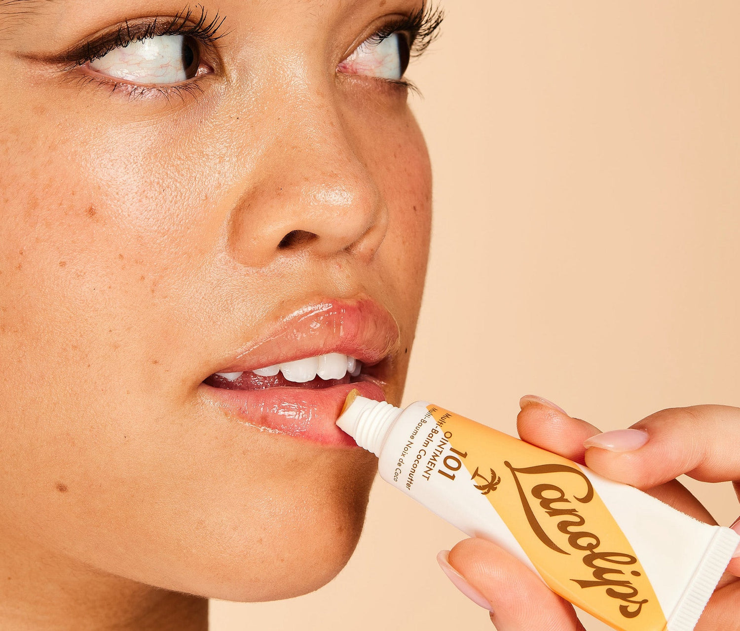 A close up shot of a model applying Lanolips 101 Ointment Multi Balm Coconutter in a caramel coloured tube. Model is pressing the applicator to her lips, looking to the side. The background is a warm peach tone, complementing the golden hues of the tube.  Using ultra-pure grade Aussie lanolin, vitamin e and coconut, his super-rich balm penetrates & seals in moisture to give long-lasting hydration for dry & chapped lips, skin patches, cuticles, elbows & more.