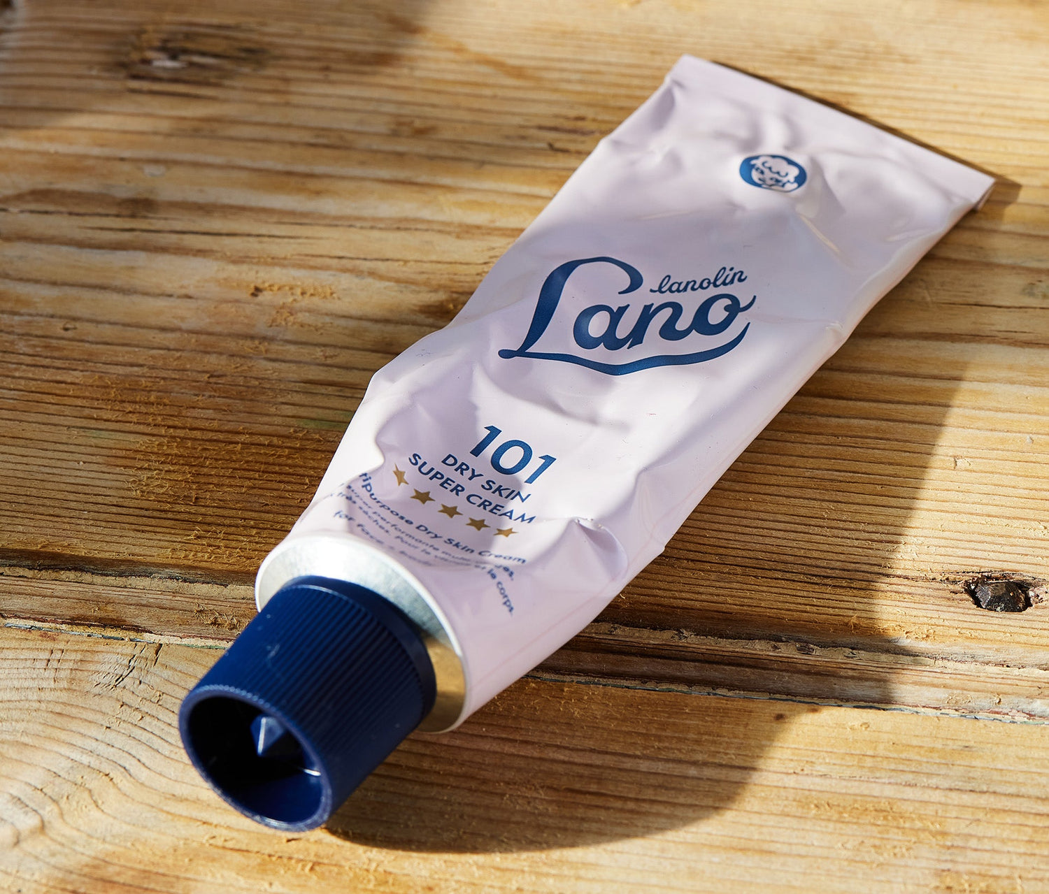 A pink tube of Lano 101 Dry Skin Super Cream rests on a wooden surface in natural sunlight. This dermatologically tested, multipurpose cream is enriched with triple lanolin, vitamin E, and shea butter to deeply hydrate and repair dry, sensitive skin. Its rich, whipped texture grips the skin before absorbing, creating a protective moisture barrier. Clinically proven to boost hydration by 98% after one use, it’s ideal for the face, hands, and body.