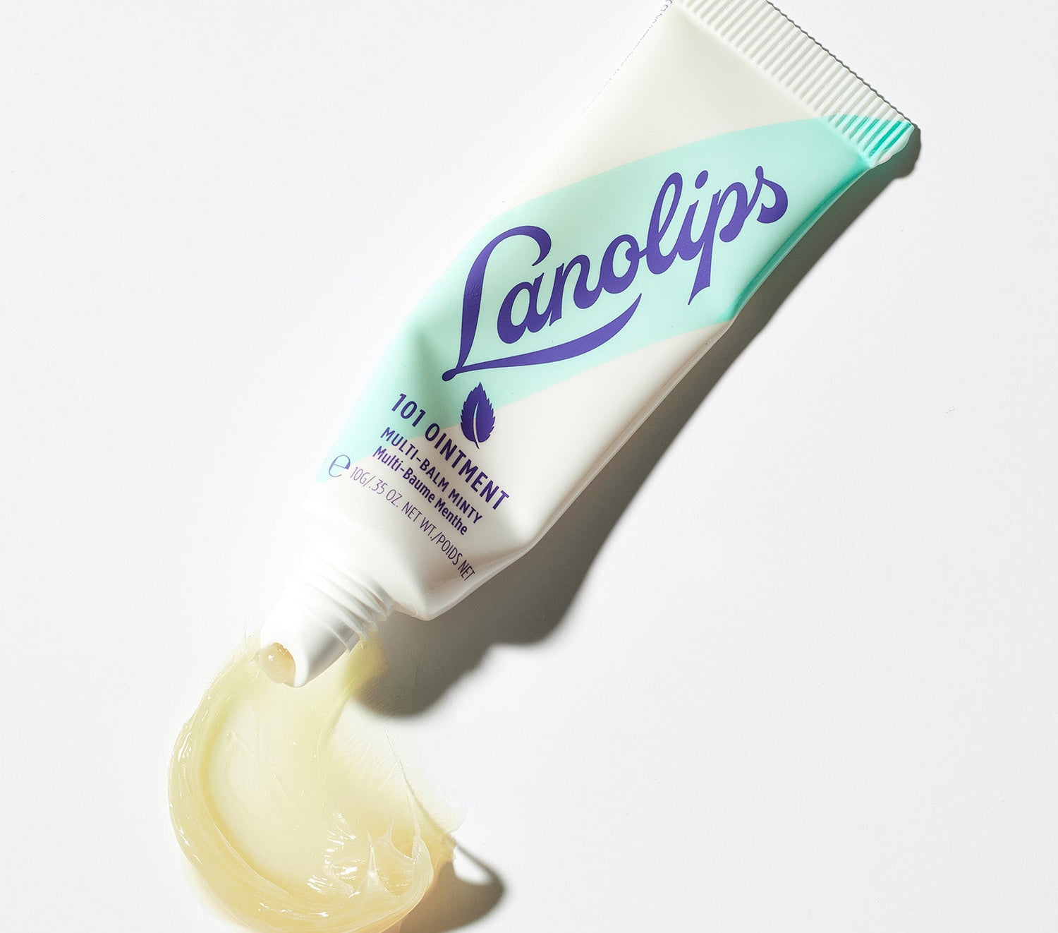 A tube of Lanolips 101 Ointment Multi-Balm in Minty is displayed on a white background, with a smooth, glossy swipe of the balm oozing out from the applicator. The tube features a refreshing mint green and white gradient with the bold blue Lanolips logo, and a small mint leaf icon. The ointment is rich, has a translucent texture. This balm contains Aussie lanolin, vitamin e, and natural peppermint oil to showcase the nourishing and soothing properties of the balm.
