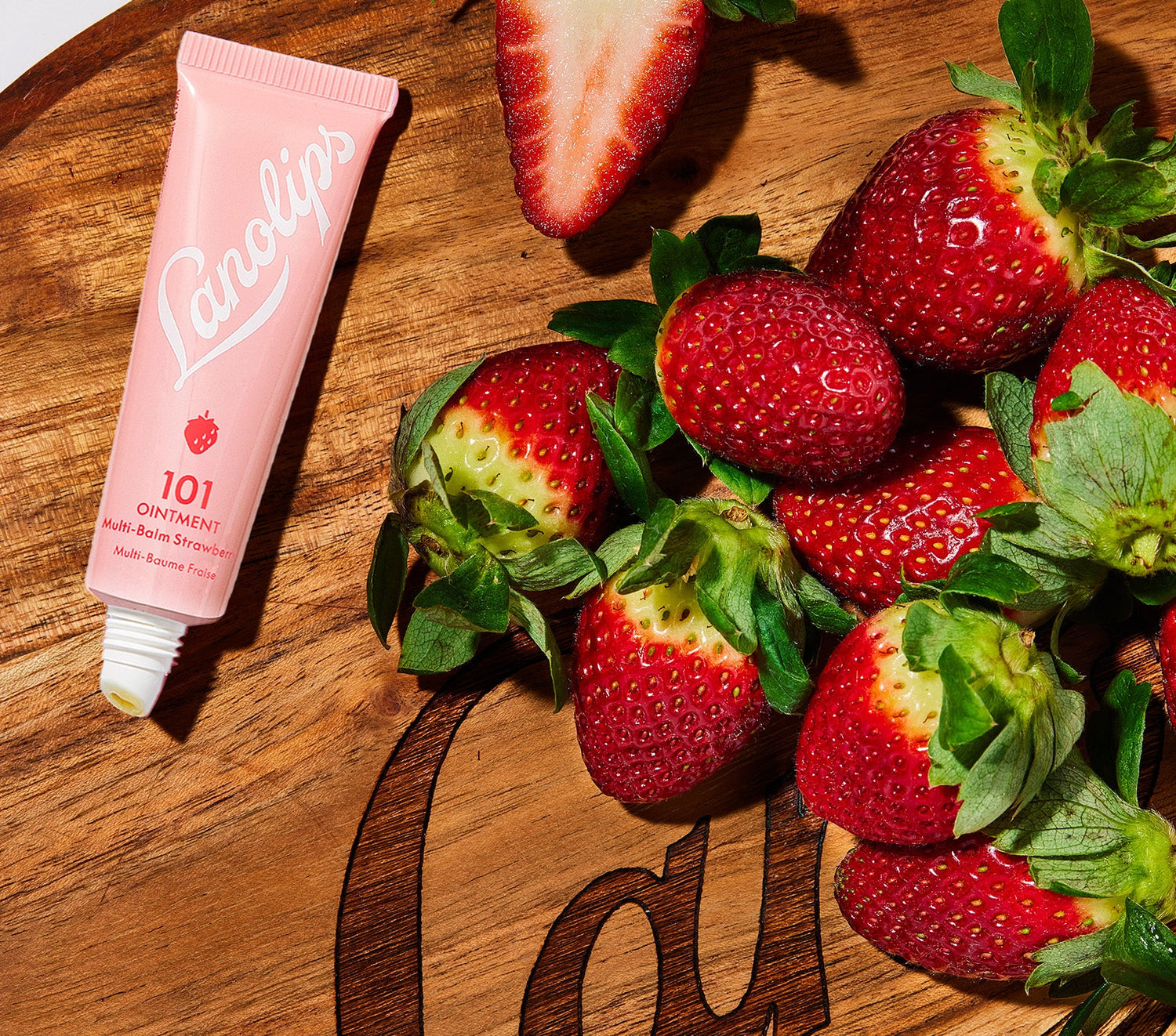 A tube of Lanolips 101 Ointment Multi Balm Strawberry, in soft pink packaging with a small strawberry illustration, placed on a wooden board next to a pile of fresh, ripe strawberries. One strawberry is sliced in half, revealing its juicy interior. Made with ultra-pure lanolin, strawberry extract, and vitamin E, this multipurpose balm provides deep hydration with a subtle fruity scent.