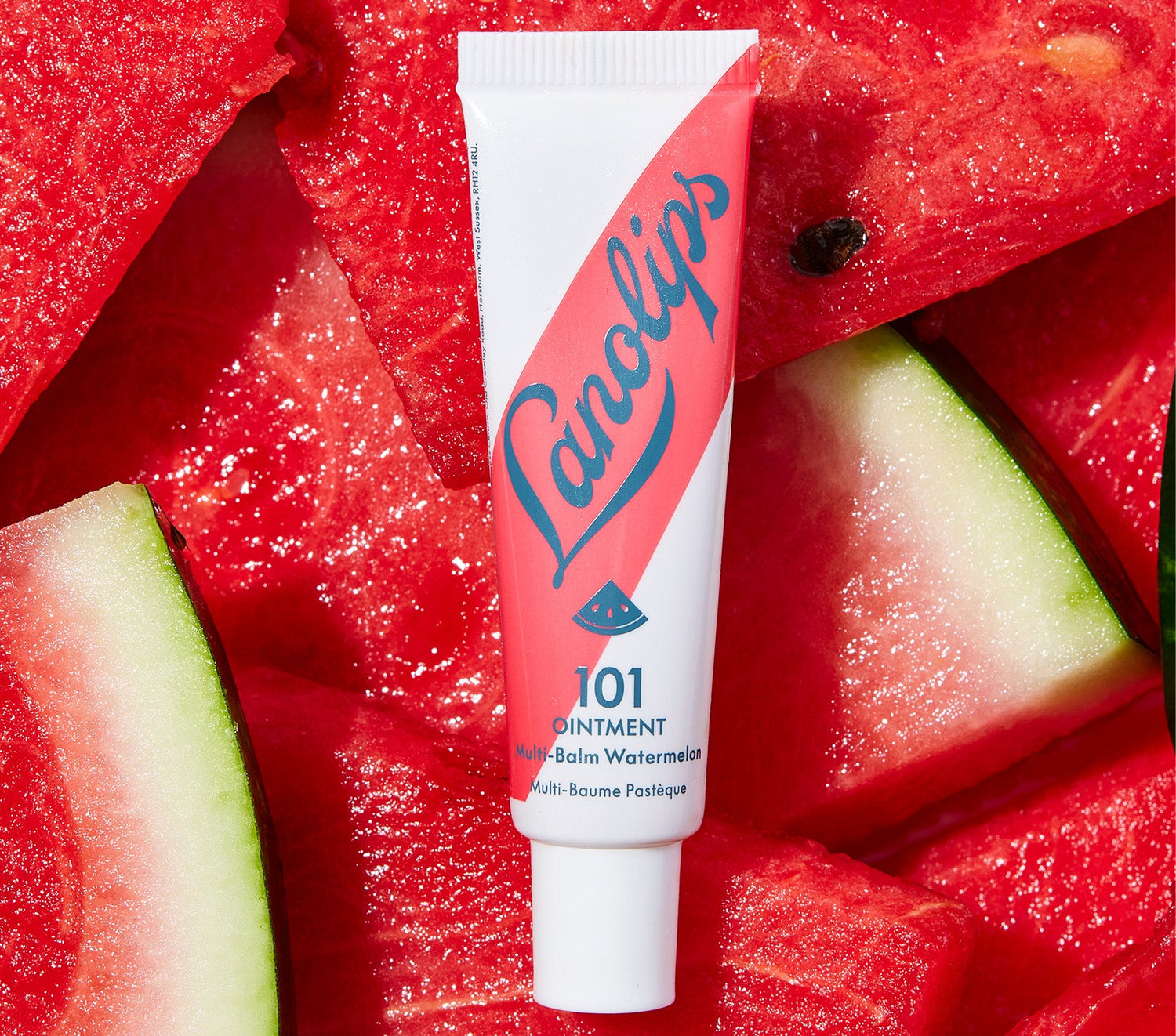 A tube of Lanolips 101 Ointment Multi Balm in Watermelon, featuring a bold pink and white design with the Lanolips blue logo and a small watermelon illustration, placed on a background of fresh, juicy watermelon pieces. Made with lanolin, vitamin e and natural watermelon fruit extract, this super rich fruity balm deeply hydrates dry and chapped lips, while delivering a refreshing scent.