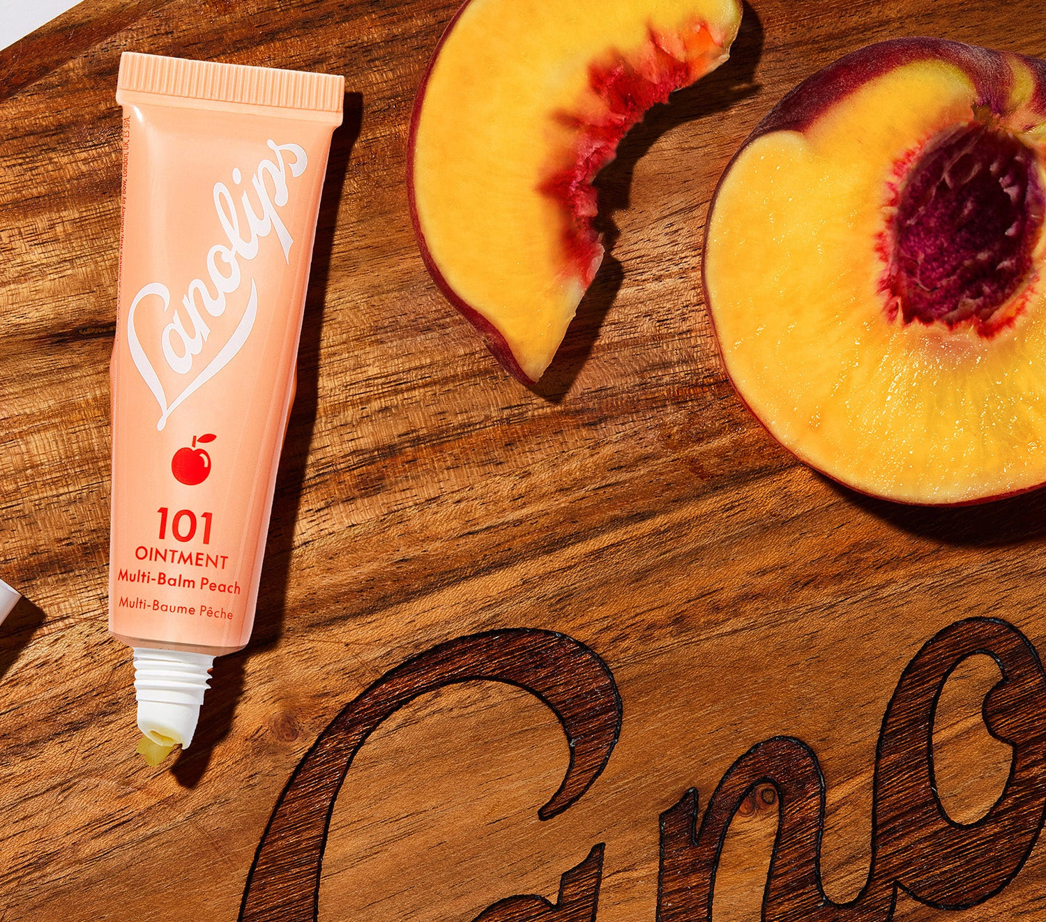 A tube of Lanolips 101 Ointment Multi Balm in Peach is placed on a wooden surface engraved with the brand's logo, alongside fresh peach slices. The tube, featuring a warm peachy orange colour with the white Lanolips logo branding and a small peach icon, is uncapped with a small amount of balm visible at the applicator top. This balm contains natural lanolin, vitamin e and peach kernel oil extracts to soften and nourish dry lips.