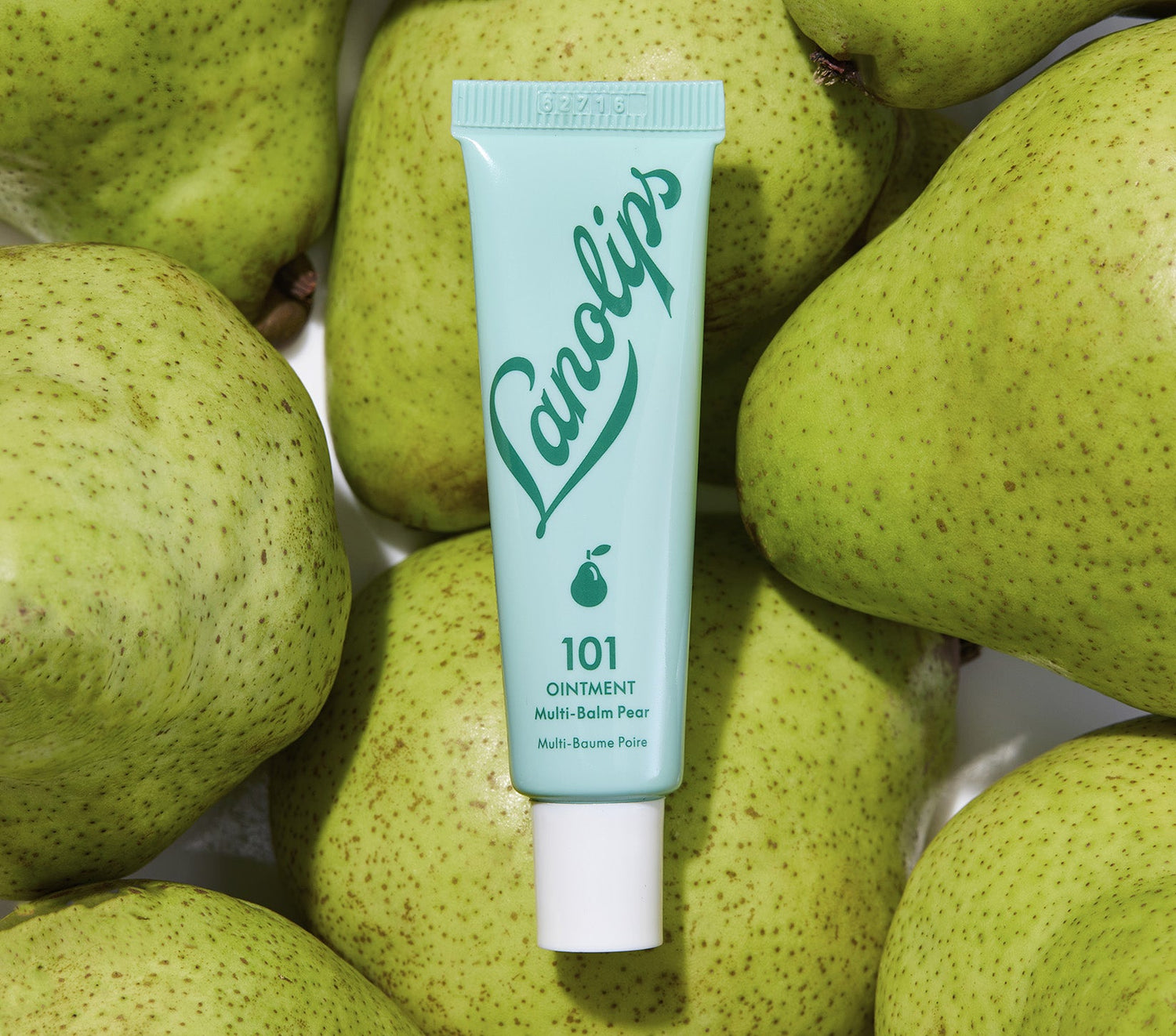 A tube of Lanolips 101 Ointment Multi Balm in Pear, in a pastel green packaging with the Lanolips logo brand, placed among fresh green pears. Made with lanolin, vitamin e and pear seed oil to give long lasting hydration for dry and chapped lips.