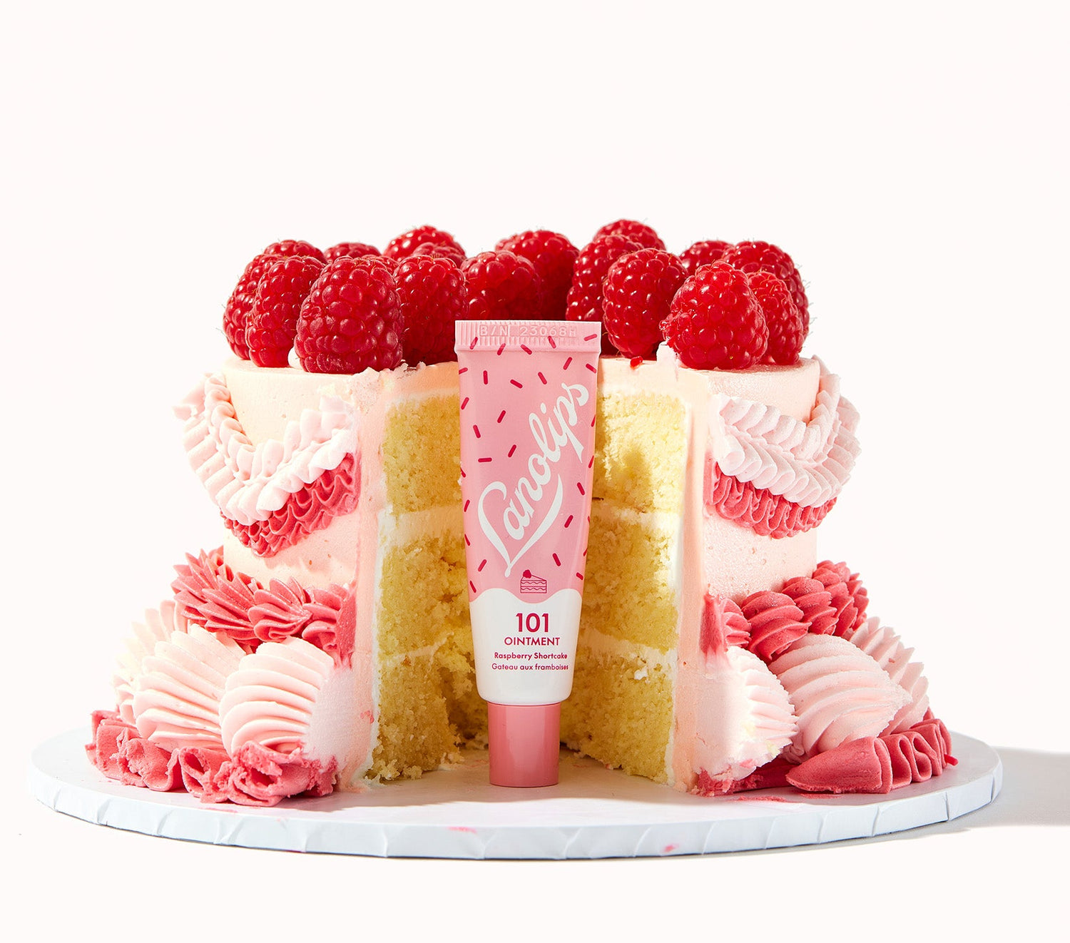A cake with pink frosting and fresh raspberries, with a tube of Lanolips 101 Ointment Multi Balm in Raspberry Shortcake placed in the centre. The balm hero ingredients include lanolin, vitamin e and all natural raspberry flavours, nourishing dry and chapped lips to a burst of sweet, tangy raspberries with rich buttery short cake goodness. Also gives long lasting hydration for dry skin patches, cuticles, elbows and more.