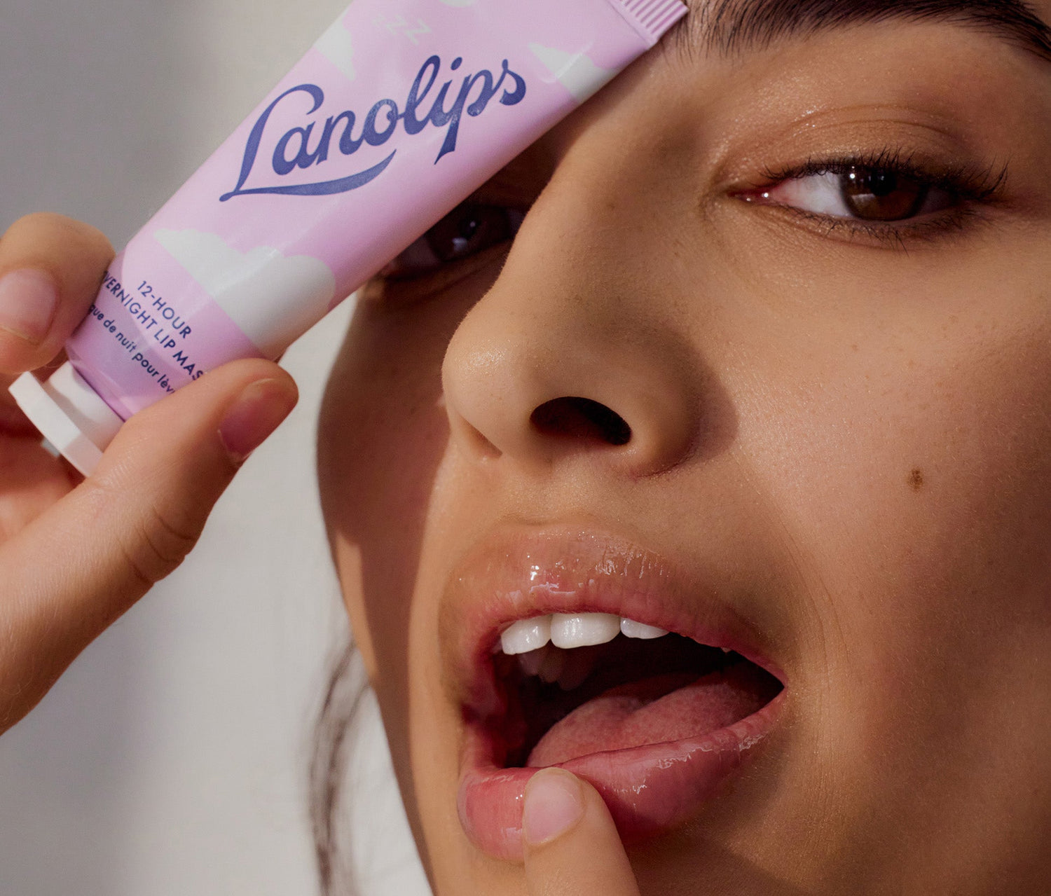A model holds a tube of Lanolips 12-Hour Overnight Lip Mask close to her face, partially covering one eye. Her lips appear glossy and deeply nourished, with a subtle sheen, as she lightly touches them with her fingertip. The lip mask contains Ultra-Pure Lanolin, which mimics the skin’s natural oils for intense overnight moisture, hyaluronic acid to plump and smooth, vitamin E for antioxidant protection, and vitamin c to soothe and repair dry lips.
