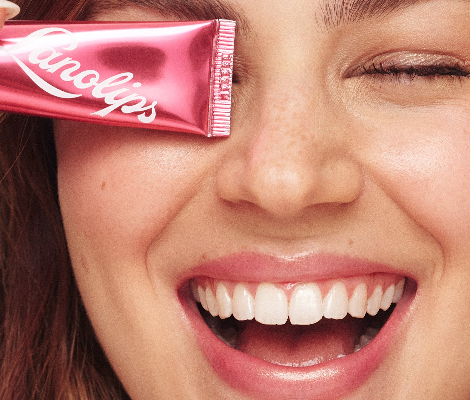A close up shot of a model wearing the Lanolips Baby Rose Gold 101 Ointment on her lips and cheeks.  The model is holding a tube, covering their left eye. their lips have a soft pink tint with a subtle sheen. Lanolips Baby Rose Gold 101 OIntment contains ultra-pure grade lanolin formula deeply moisturises while natural oils and waxes lock in hydration, leaving the lips looking plump and nourished. The model’s complexion appears luminous, with a soft golden highlight on her cheekbones.