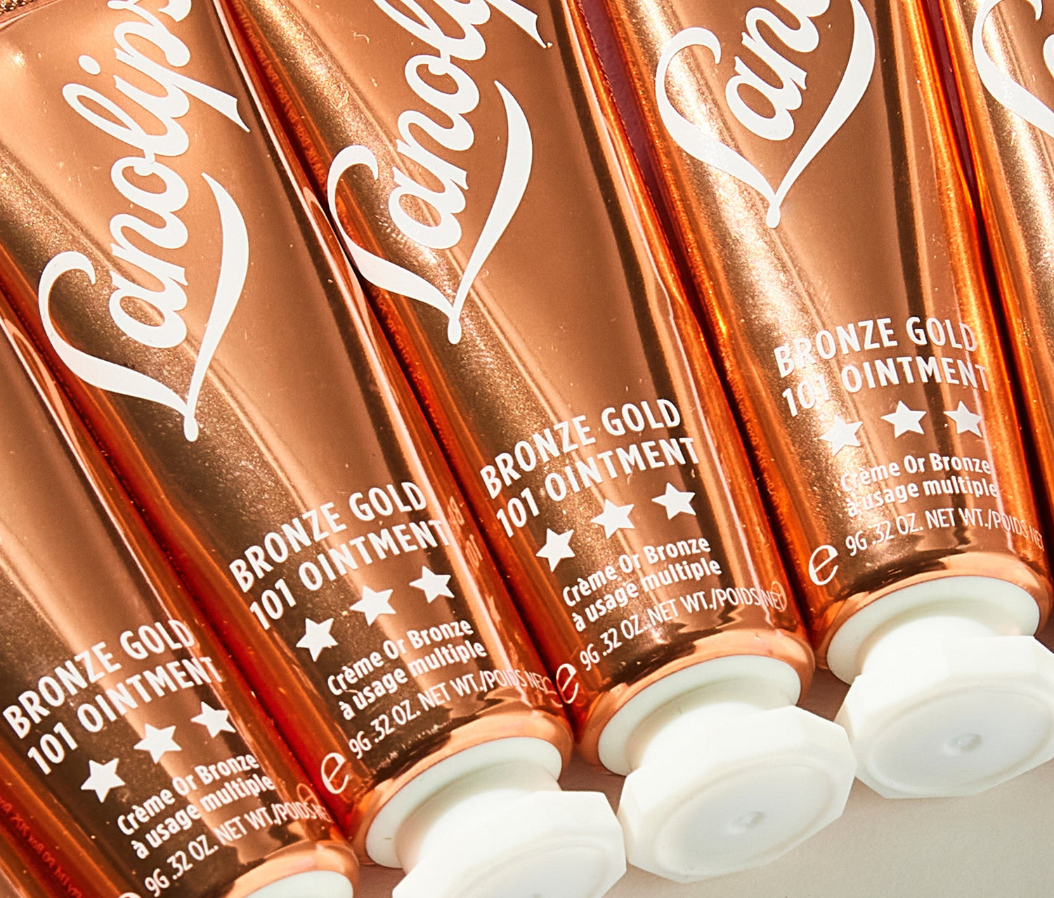 A close-up of multiple tubes of Lanolips Bronze Gold 101 Ointment, arranged in a diagonal pattern. This ointment contains Ultra-Pure Lanolin, which mimics the skin’s natural oils for deep hydration, along with subtle bronze-gold pigments to provide a sheer, sun-kissed glow. Infused with natural oils and waxes, the balm locks in moisture while delivering a soft, adiant shimmer to lips and skin. The composition highlights the luxurious, nourishing, and illuminating benefits of the ointment.