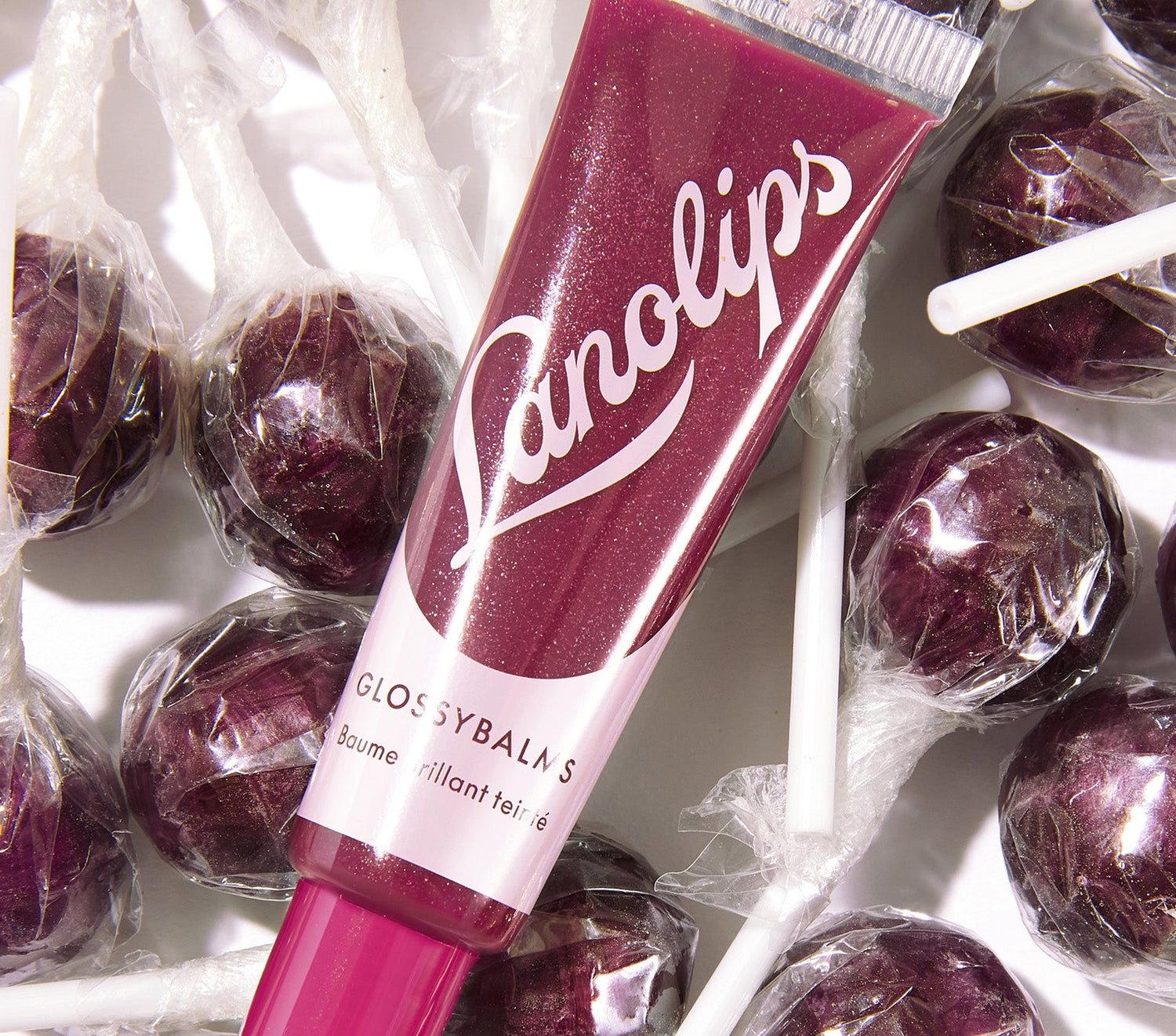 A tube of Glossy Balm Berry in a deep berry shade with a shimmering finish, featuring bold light pink Lanolips logo. The balm is surrounded by wrapped purple lollipops, matching its rich hue. Enriched with all-natural lanolin that smooths over dryness, seals in moisture and drenches lips in non-sticky hydration that lasts for hours.