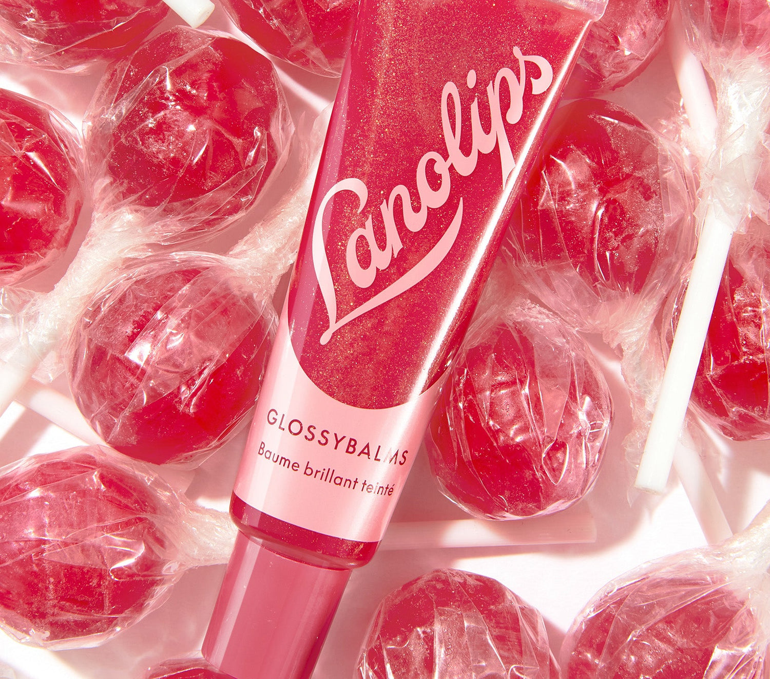 A tube of Glossy Balm Candy in a fruity pink shade with a shimmering finish, featuring bold light pink Lanolips logo. The balm is surrounded by wrapped pink lollipops, matching its bright hue. Enriched with all-natural lanolin that smooths over dryness, seals in moisture and drenches lips in non-sticky hydration that lasts for hours.