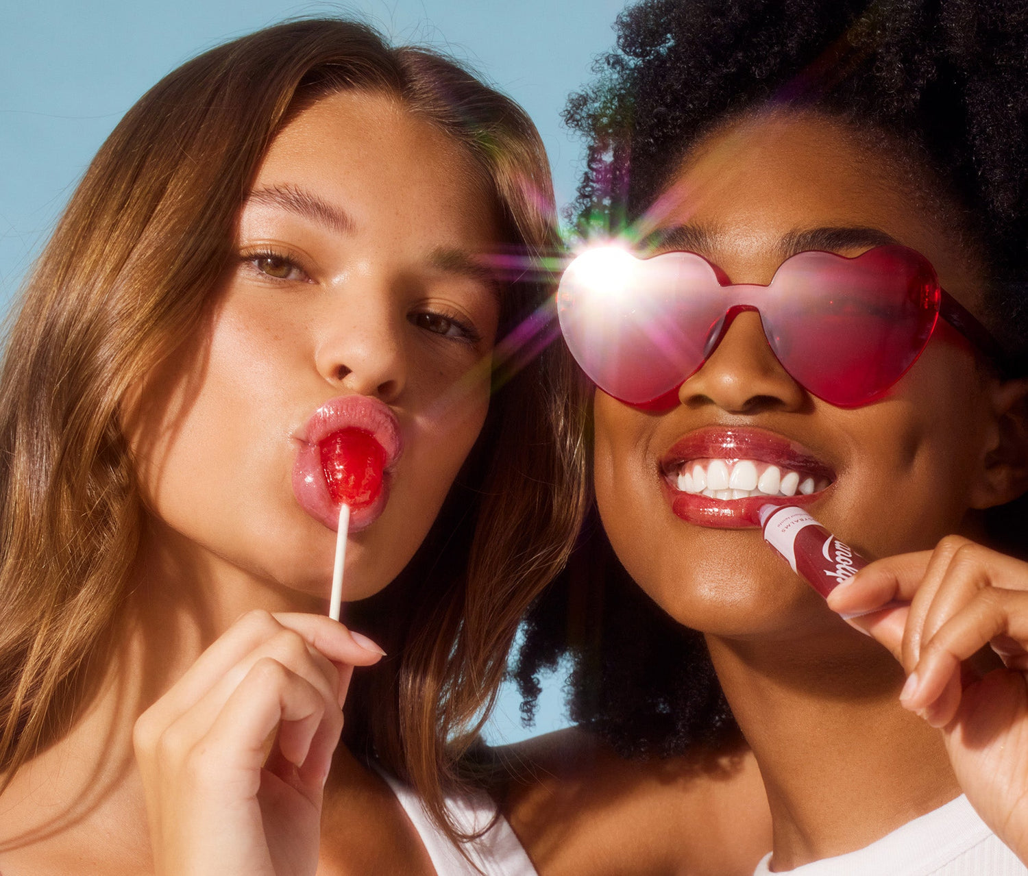 Two models posed closely together against a light blue background. One model, with wavy brown hair, puckers her lips while licking a red lollipop. The other, with voluminous curls and red heart-shaped sunglasses reflecting bright light, smiles while holding a tube of Lanolips Glossy Balm in a deep berry shade. The glossy lip balm is made of all-natural lanolin that smooths over dryness, seals in moisture and drenches lips in non-sticky hydration that lasts for hours.