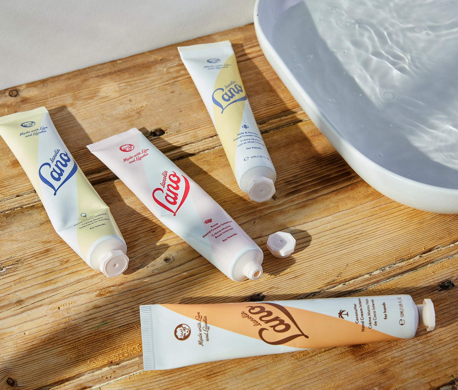 Four tubes of Lanolips Hand Cream Intense rest on a rustic wooden surface beside a bowl of water. Infused with triple lanolin, this rich hand cream deeply nourishes and repairs dry skin, creating a protective moisture barrier. The collection features four indulgent flavours - Coconutter, Rose, Milk & Honey, and Vanilla—each offering long-lasting hydration with a luxurious feel. Perfect for soft, smooth hands, this dermatologically tested formula absorbs beautifully without feeling greasy.