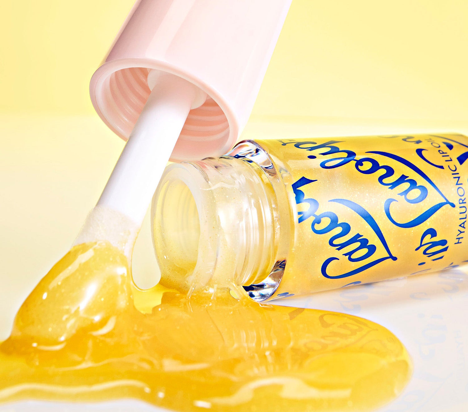 A close up shot of an open tube of Lanolips Hyaluronic Lip Oil in Honey, with golden, glossy lip oil spilling onto a smooth surface. The clear tube features elegant blue lettering, and the soft pink cap holds a doe-foot applicator coated in the rich, honey-infused formula. Made with ultra-pure lanolin, hyaluronic acid, and vitamin e, this lip oil delivers intense hydration, a golden shimmer, and a lightweight, non-sticky finish for soft, radiant lips.