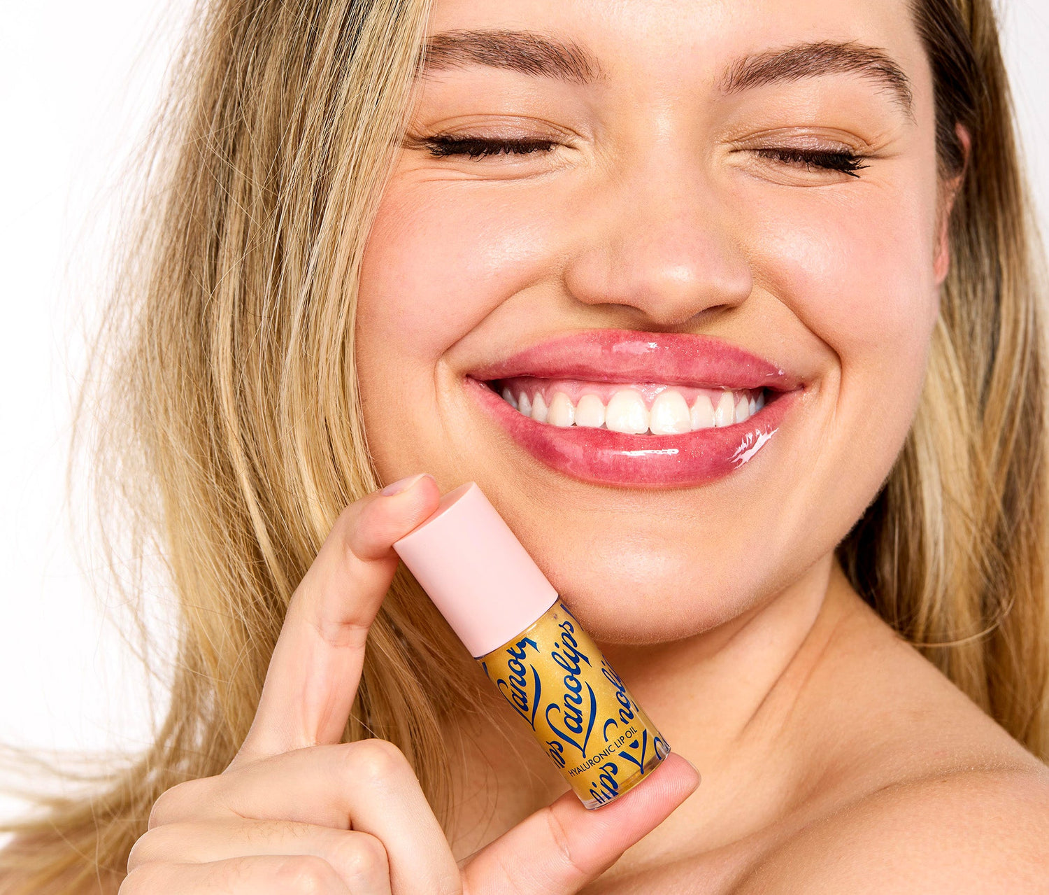 A model holds a tube of Lanolips Hyaluronic Lip Oil in Honey close to her face. The lip oil is in a soft beige and pink lid with the tube having the Lanolips logo wrapped around it. Made with ultra-pure lanolin, hyaluronic acid, and vitamin e, this lip oil delivers intense hydration, a golden shimmer, and a lightweight, non-sticky finish for soft, radiant lips.