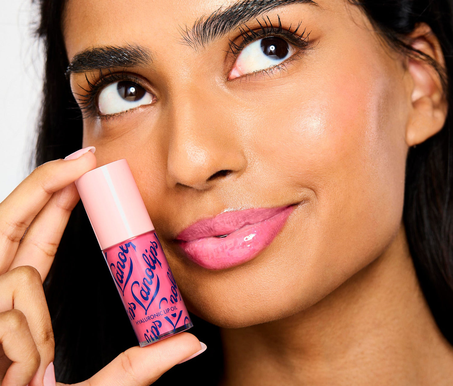 A model holds a tube of Lanolips Hyaluronic Lip Oil Raspberry close to her face. The Hyaluronic Lip Oil is in a tube with bold Lanolips lettering, topped with a soft pink lid. The model is wearing the lip oil on her lips, showing the sheer raspberry tint. Made with ultra rich lanolin, hyaluronic acid and vitamin e, this lip oil delivers intense hydration, with fruity pink tint that adds a natural flush of colour to your lips.