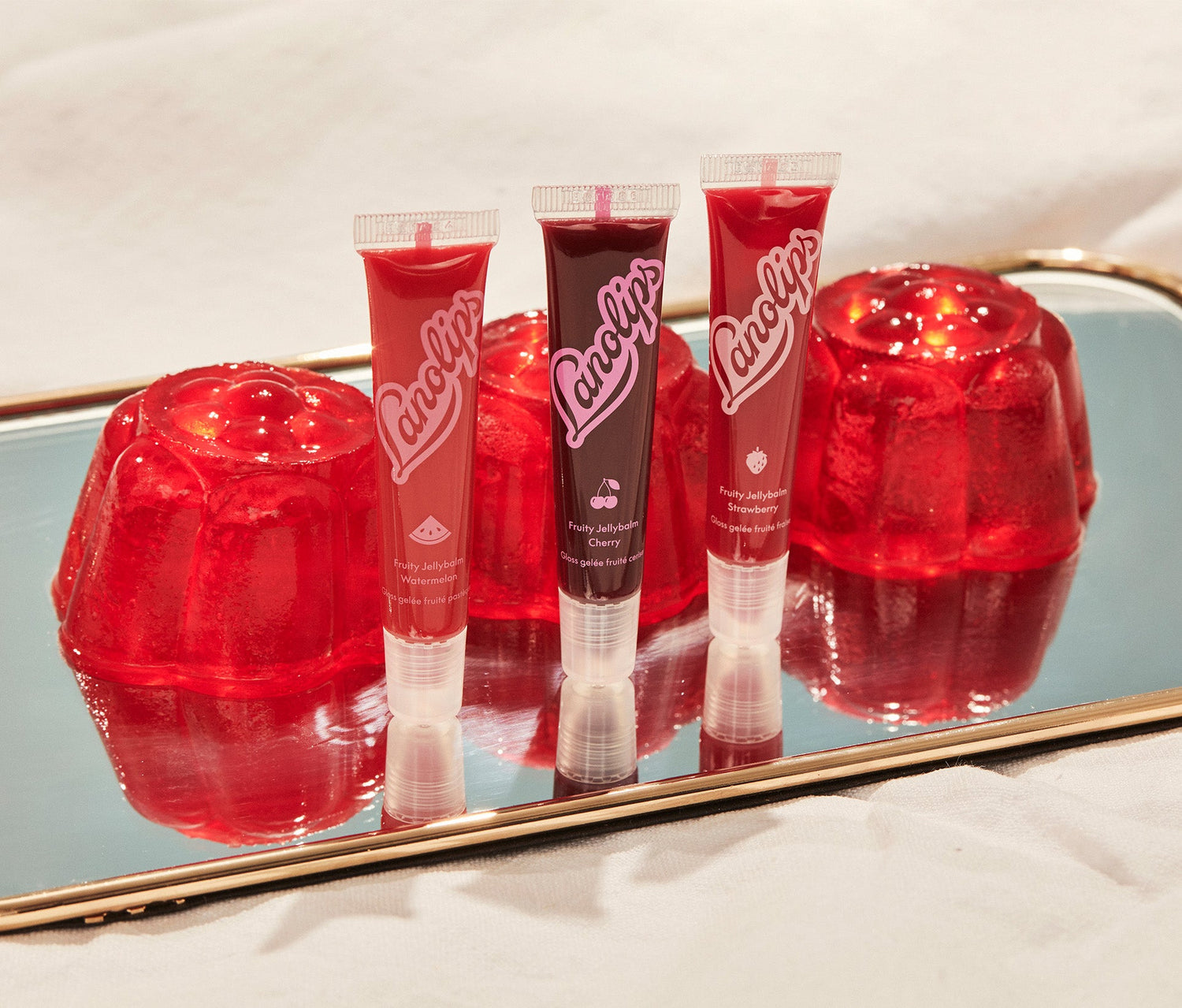 The Lanolips Jellybalm range comes in three fruity flavours: Watermelon, Cherry, and Strawberry. The tubes are standing upright on a reflective gold-trimmed mirror, surrounded by glossy red jelly moulds. The vibrant red and pink packaging features fruit icons, highlighting the juicy, hydrating formulas. Made with ultra-rich lanolin and all-natural fruit extracts, these balms deliver deep hydration with a sheer, glossy finish inspired by fresh, fruity flavours.
