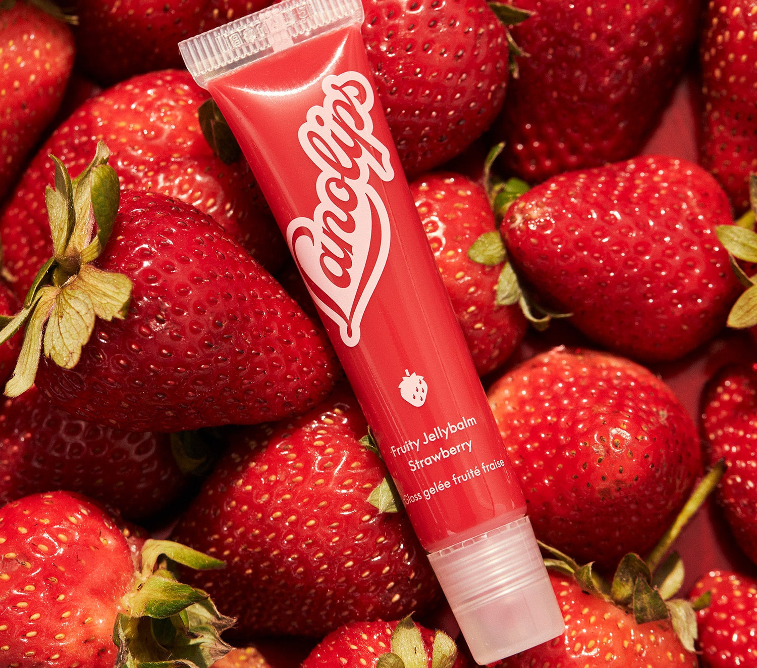 A tube of Lanolips Fruity Jellybalm in Strawberry, placed among fresh, ripe strawberries. The vibrant red packaging features the Lanolips logo and a small strawberry icon, highlighting its juicy, fruit-inspired formula. Made ultra-rich lanolin and all natural strawberry extract, this balm delivers deep moisture and a sheer, glossy tint inspired by the sweetness of fresh strawberries.