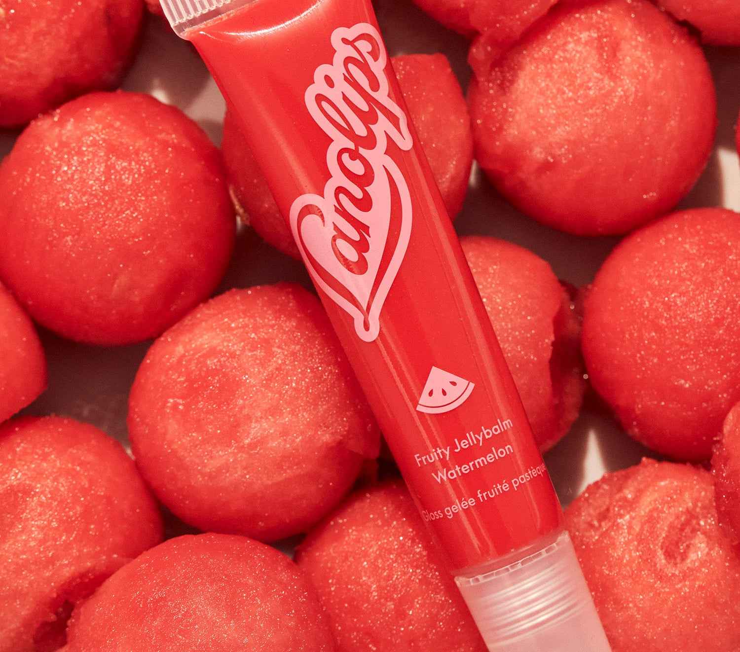 A tube of Lanolips Jellybalm Watermelon is placed on top of watermelon balls. The texture glossy red and the tube has the Lanolips logo in pink. Made with ultra rich lanolin, all natural watermelon fruit extract, and hydrating jelly for soft and glossy lips.