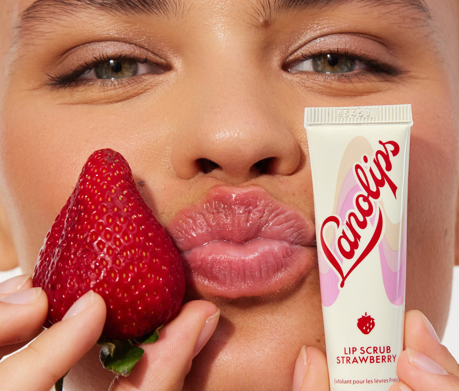 A model puckers her lips while holding a ripe strawberry in one hand and a tube of Lanolips Lip Scrub Strawberry in the other. Lanolips’ Lip Scrub Strawberry is a 100% natural balm-based scrub made with gentle sugar and real, finely ground strawberry seeds to smooth away dry, flaky skin. The formula is enriched with 70% lanolin, instantly leaving lips soft and supple while revealing a healthy lip colour and natural glow.