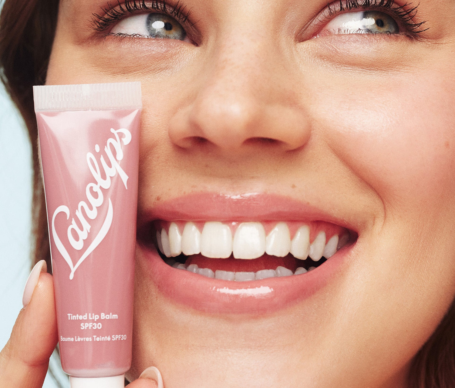 A model holding a tube of Lanolips Tinted Lip Balm SPF30 Perfect Nude close to her face. Their lips have a sheer, creamy nude tint with a soft, hydrated glow. Made with lanolin, shea butter, vitamin e and SPF30, this lanolin rich lip moisturiser deeply nourishes while protecting against sun damage. The sheer colour tint suits all skin tones, giving a natural effortless look - perfect for casual outings  or weekend coffee runs.