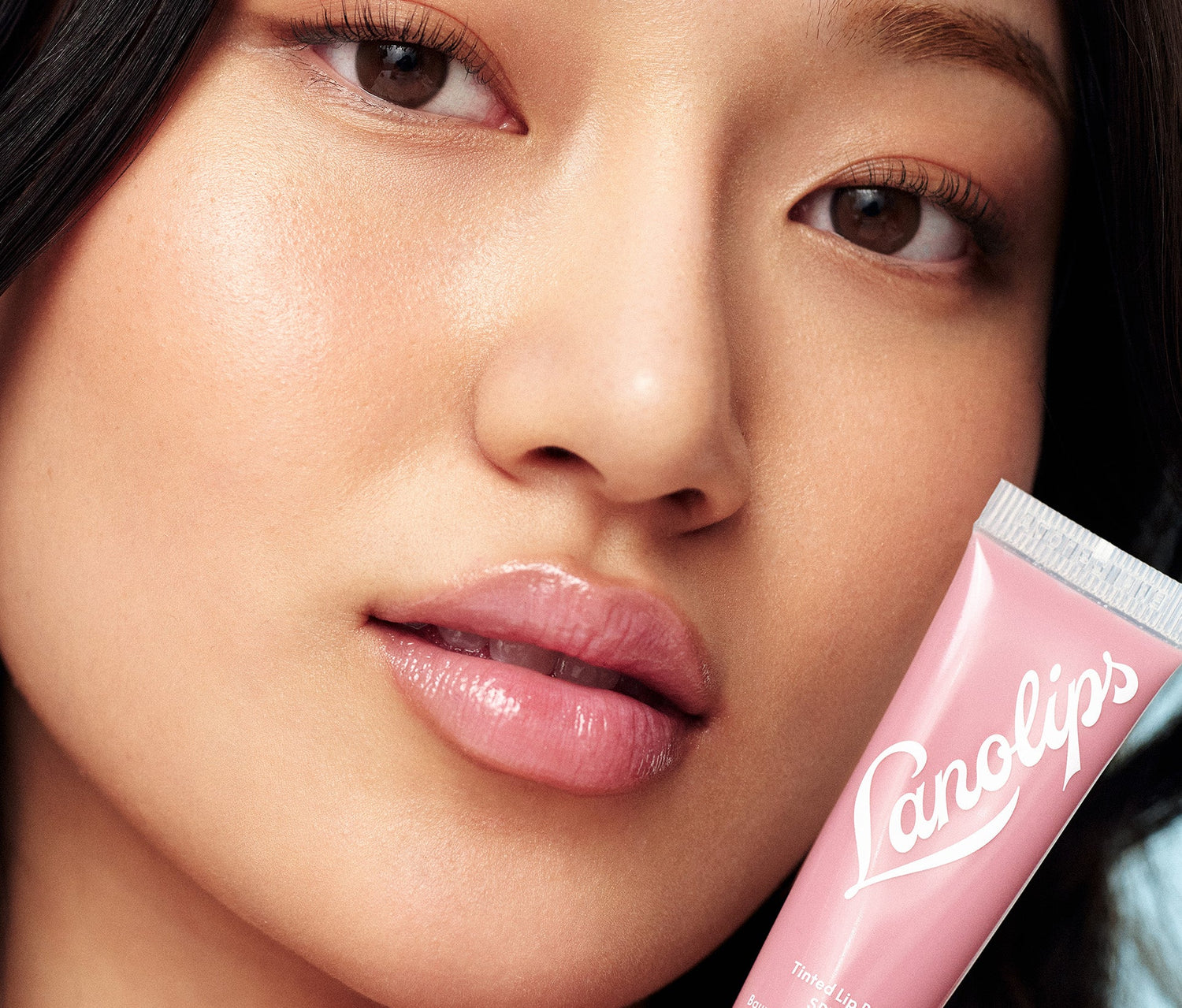 Close up shot of a model wearing a sheer colour tint from a lanolin-rich lip moisturiser with SPF30. She holds a tube of Lanolips Tinted Lip Balm SPF30 in Rose, formulated with hydrating lanolin, vitamin E, and shea butter for soft, nourished lips. The SPF30 protection helps shield against sun damage while providing a natural, dewy finish.