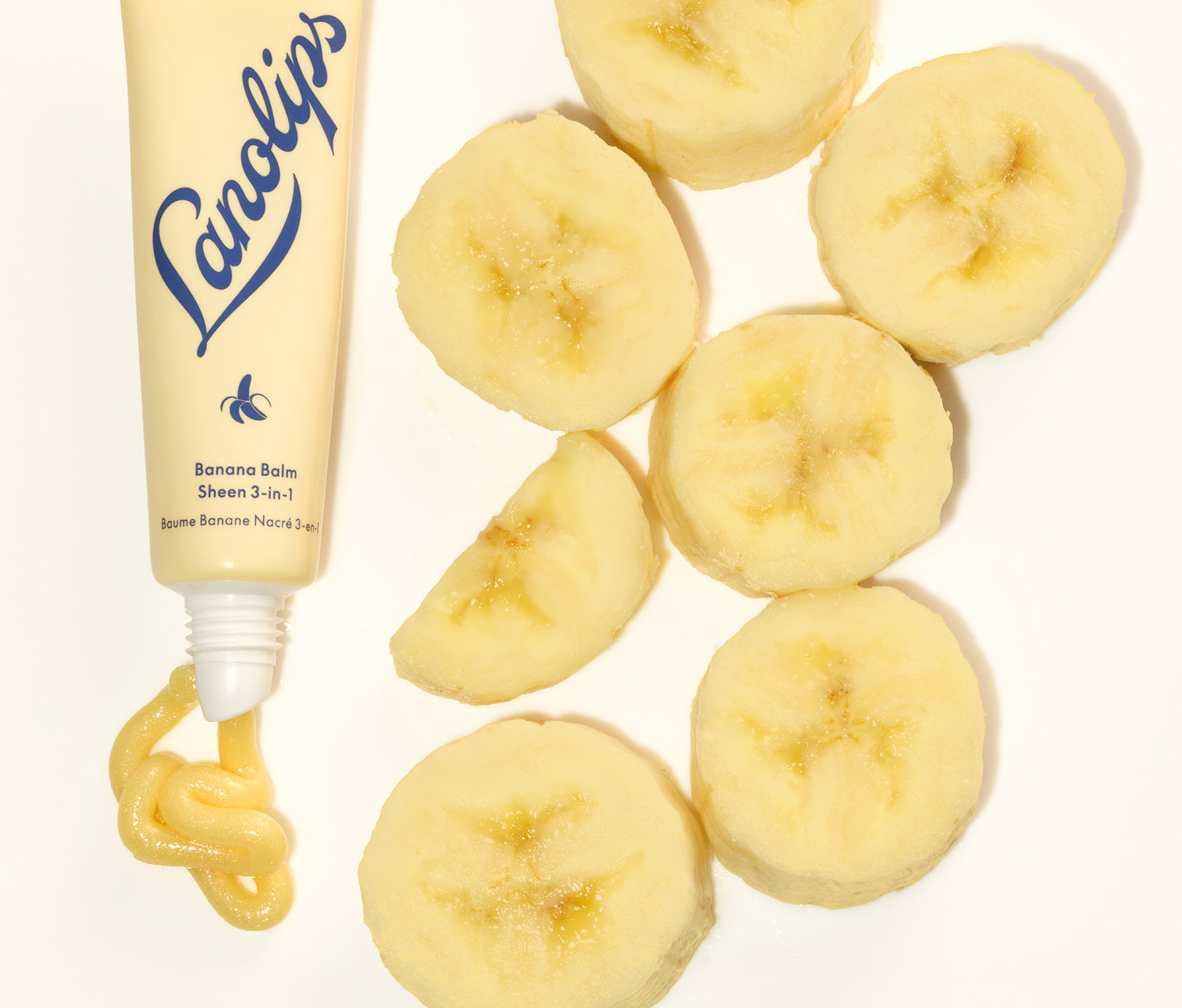 A squeezed tube of Lanolips Banana Balm 3-in-1 Lip Sheen, with fresh bananas placed on the side. Made with lanolin, banana extract, cocoa seed butter and a hint of shimmer, this balm nourishes while delivering a soft, fruity scent.
