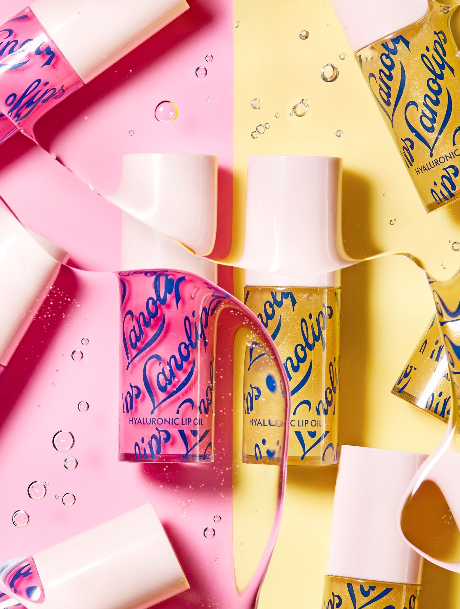 Lifestyle image of Lanolips Hyaluronic Lip Oils in Honey and Raspberry shades, arranged on a pink and yellow split background with oil droplets and flowing lip oil creating a dynamic composition