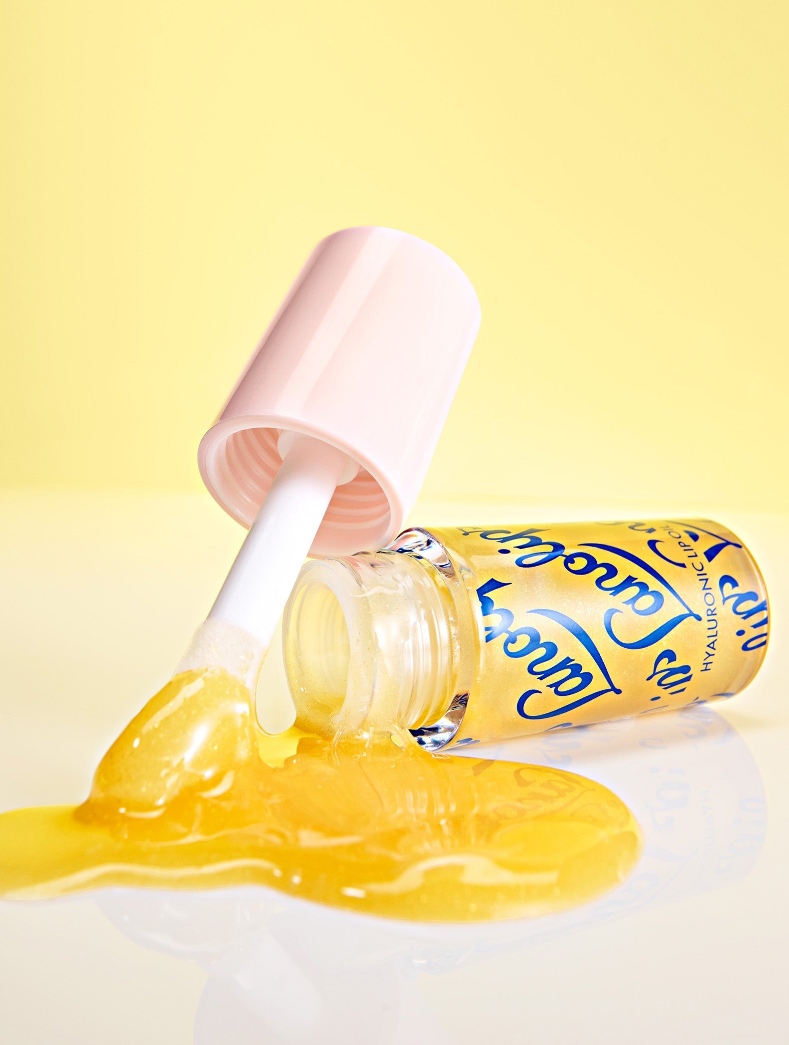 Lifestyle image of Lanolips Hyaluronic Lip Oil in the shade Honey, arranged on a yellow background showing the lip oil droplets and lip oil flowing from the container for a dynamic, glossy effect.